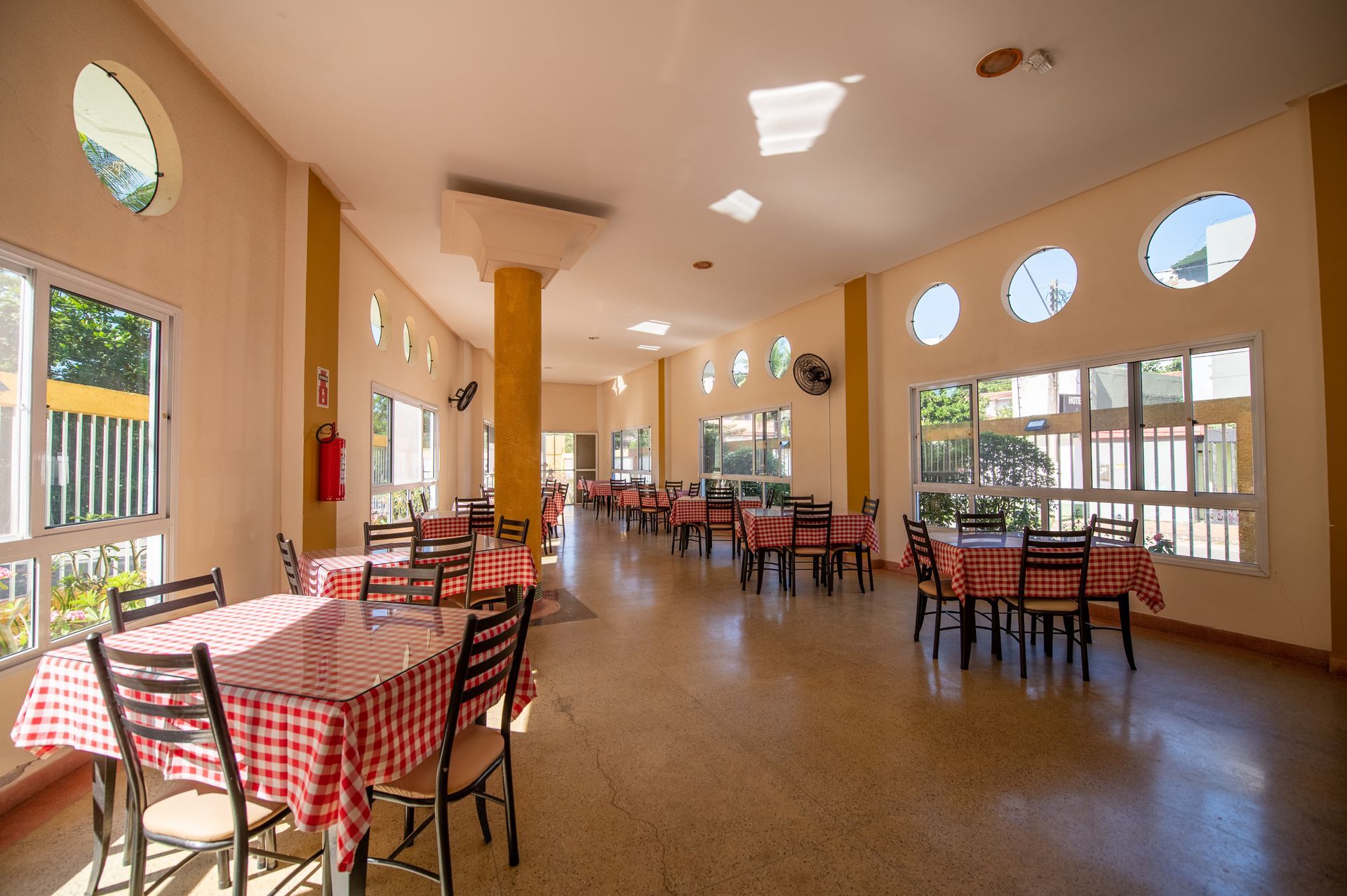 Sala de jantar de restaurante iluminada com mesas e cadeiras xadrez vermelhas e grandes janelas.