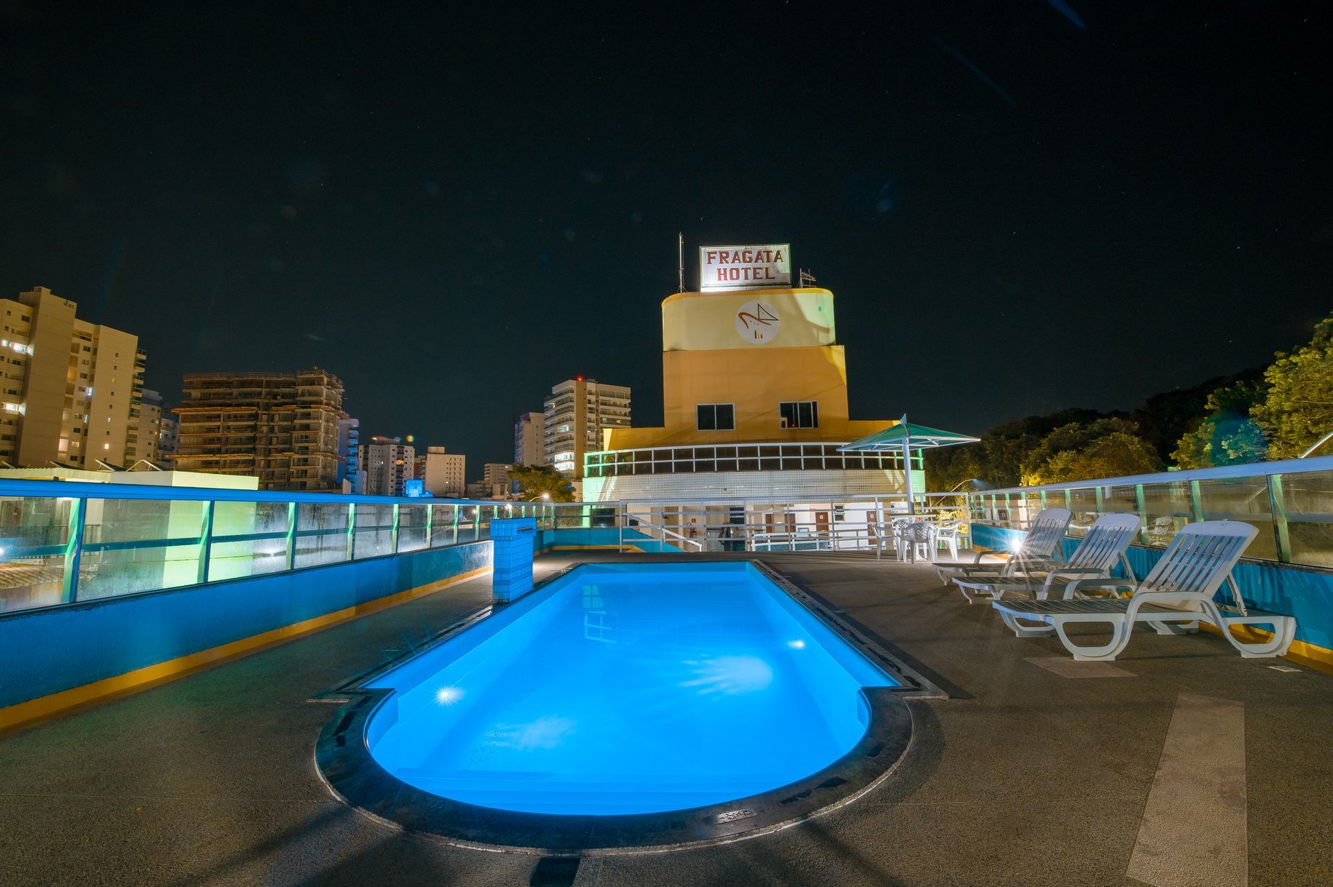 Piscina noturna na cobertura com água azul brilhante, espreguiçadeiras, horizonte da cidade e prédios iluminados ao fundo.