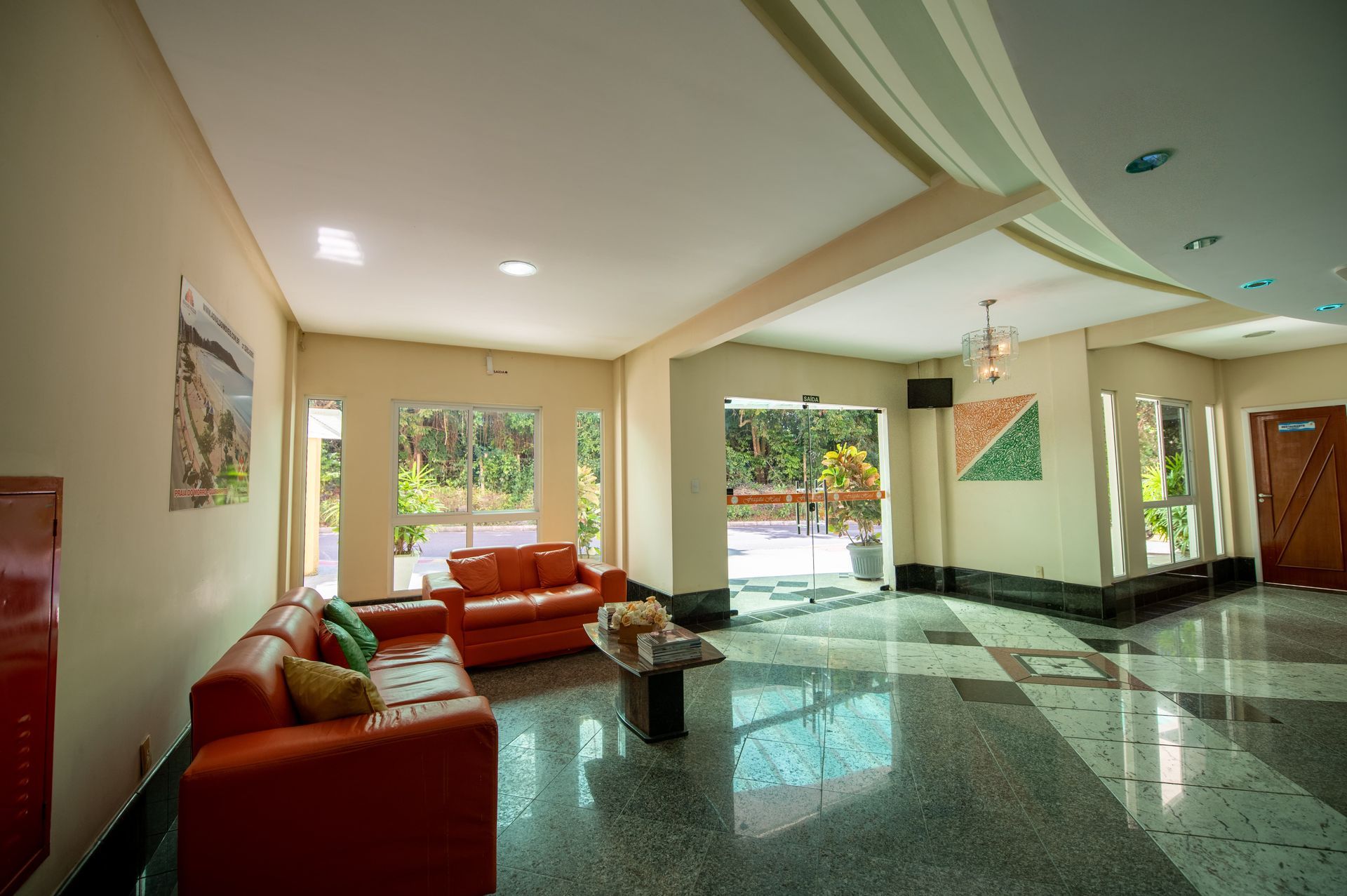 Hall de entrada moderno do hotel com sofás laranja, pisos brilhantes e grandes janelas com vista para a vegetação.
