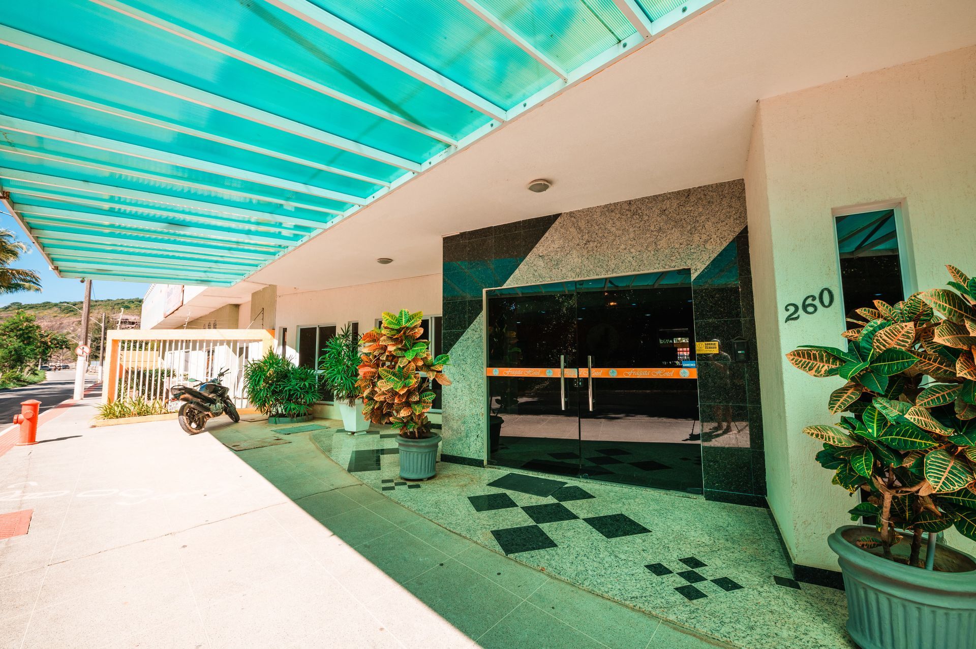 Entrada externa do motel com toldo turquesa, vasos de plantas e carro estacionado perto do quarto 280.