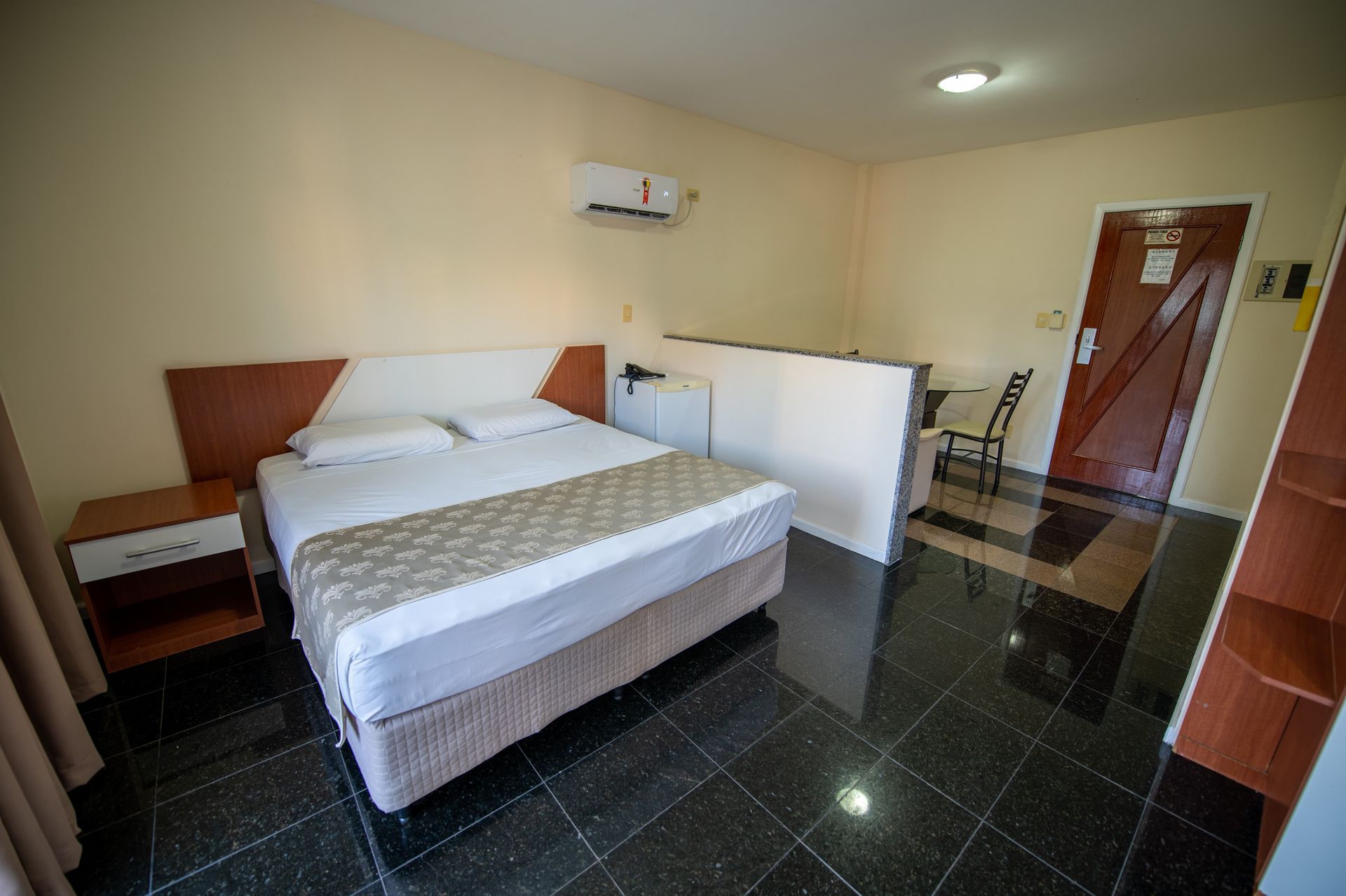 Quarto de hotel simples com cama, mesa de cabeceira, piso de azulejo preto e uma pequena escrivaninha junto à janela.