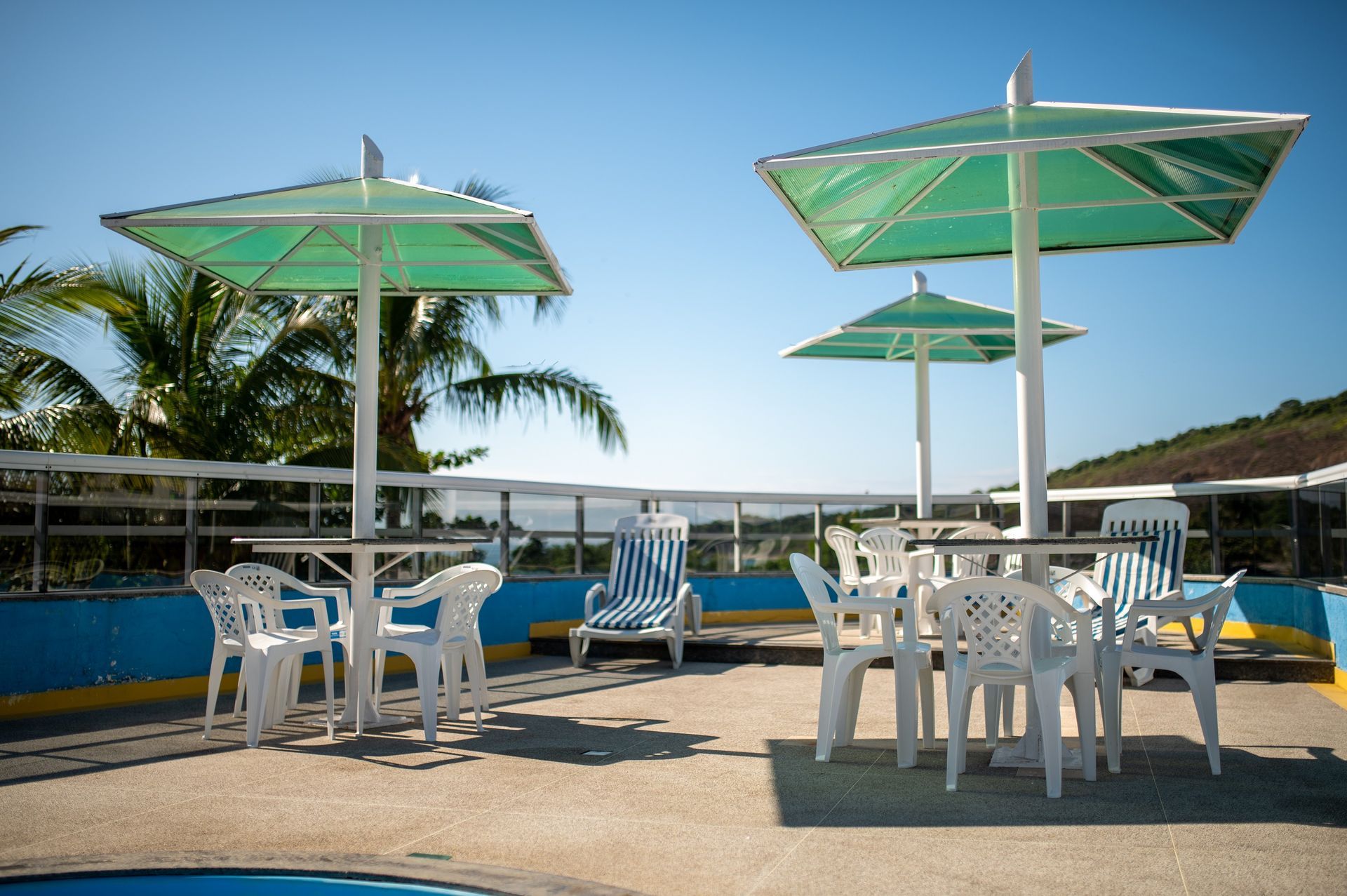 Pátio à beira da piscina com mesas e cadeiras brancas sob guarda-sóis verdes, ao lado de palmeiras e água azul.
