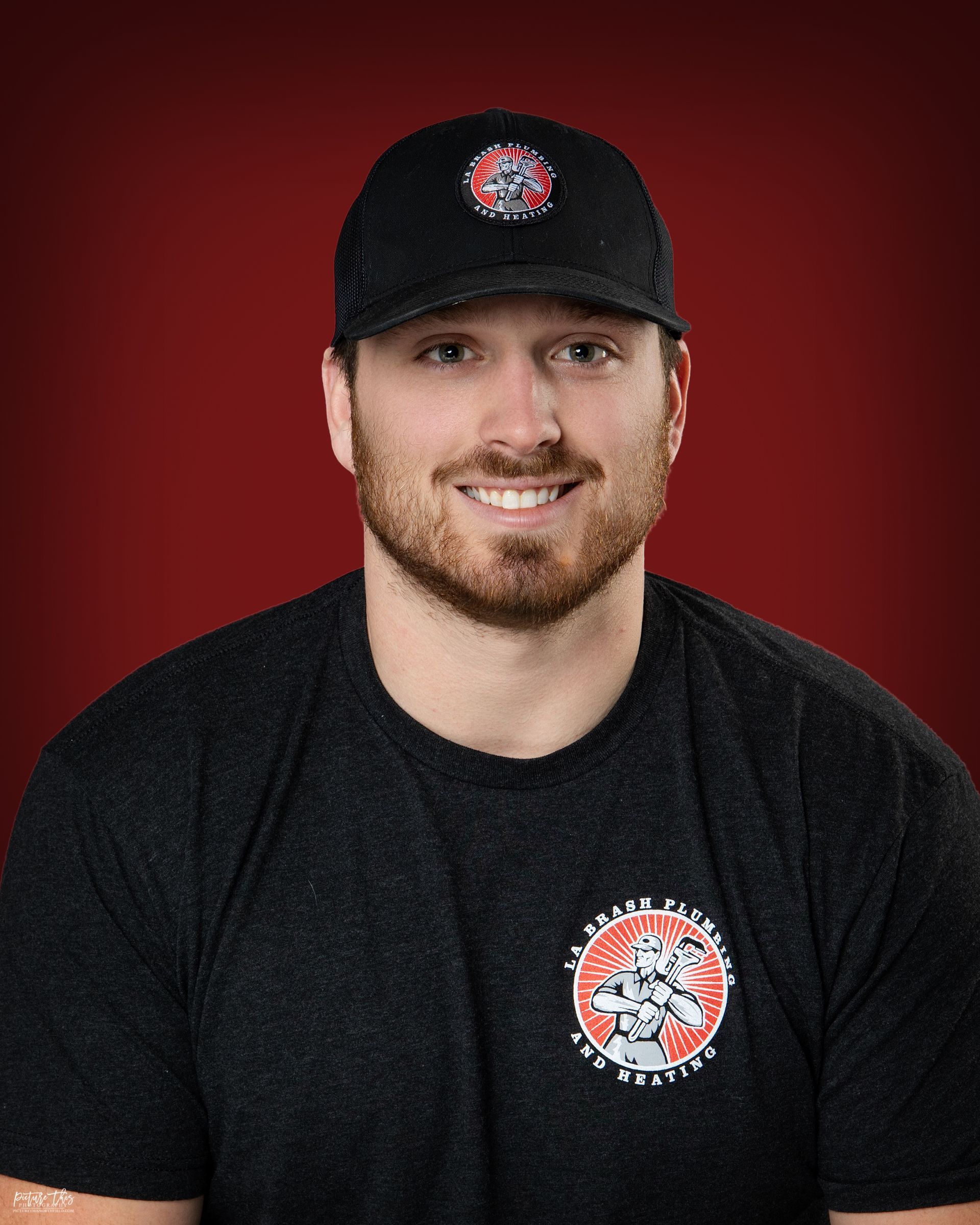 Man with a beard wearing a black cap and shirt smiles, with a red background.