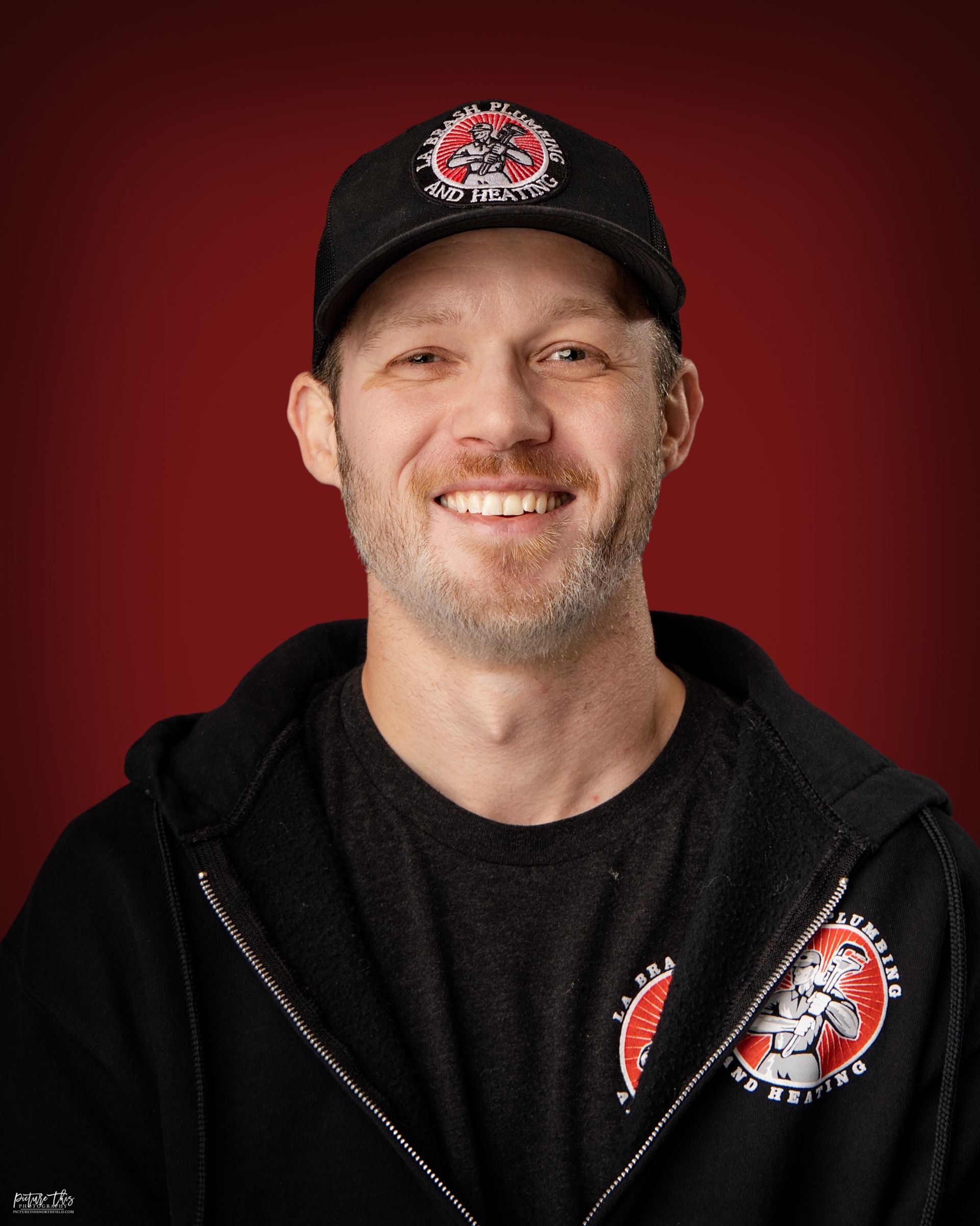 Man smiling, wearing a black cap and zip-up hoodie with a logo. Red background.
