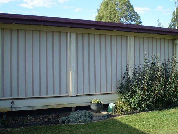 Beige corrugated metal siding on a building with a brown roof, green grass, and shrubbery.