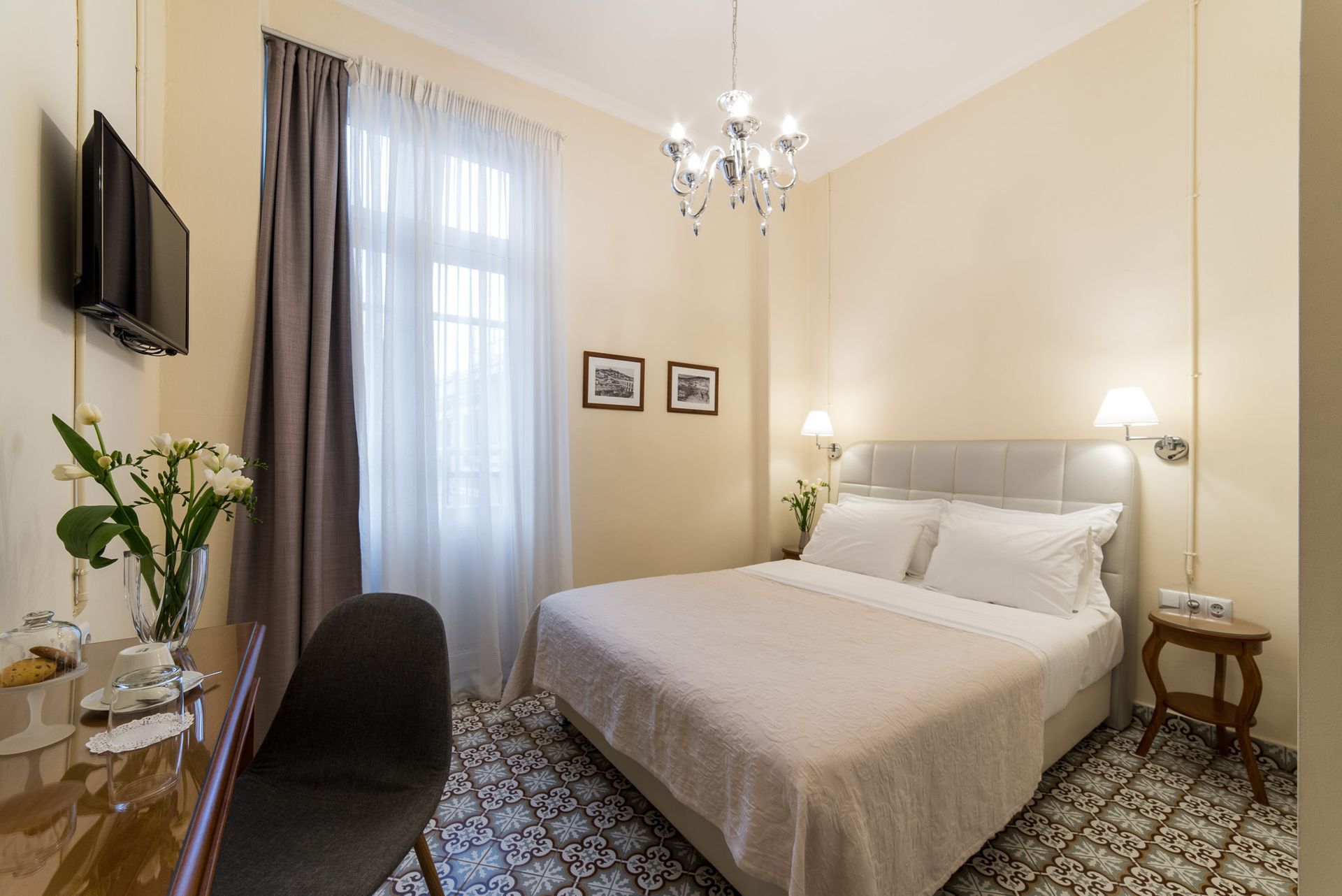 A cozy bedroom with a queen bed, crystal chandelier, decorative tile flooring, a desk, and a wall-mounted TV.
