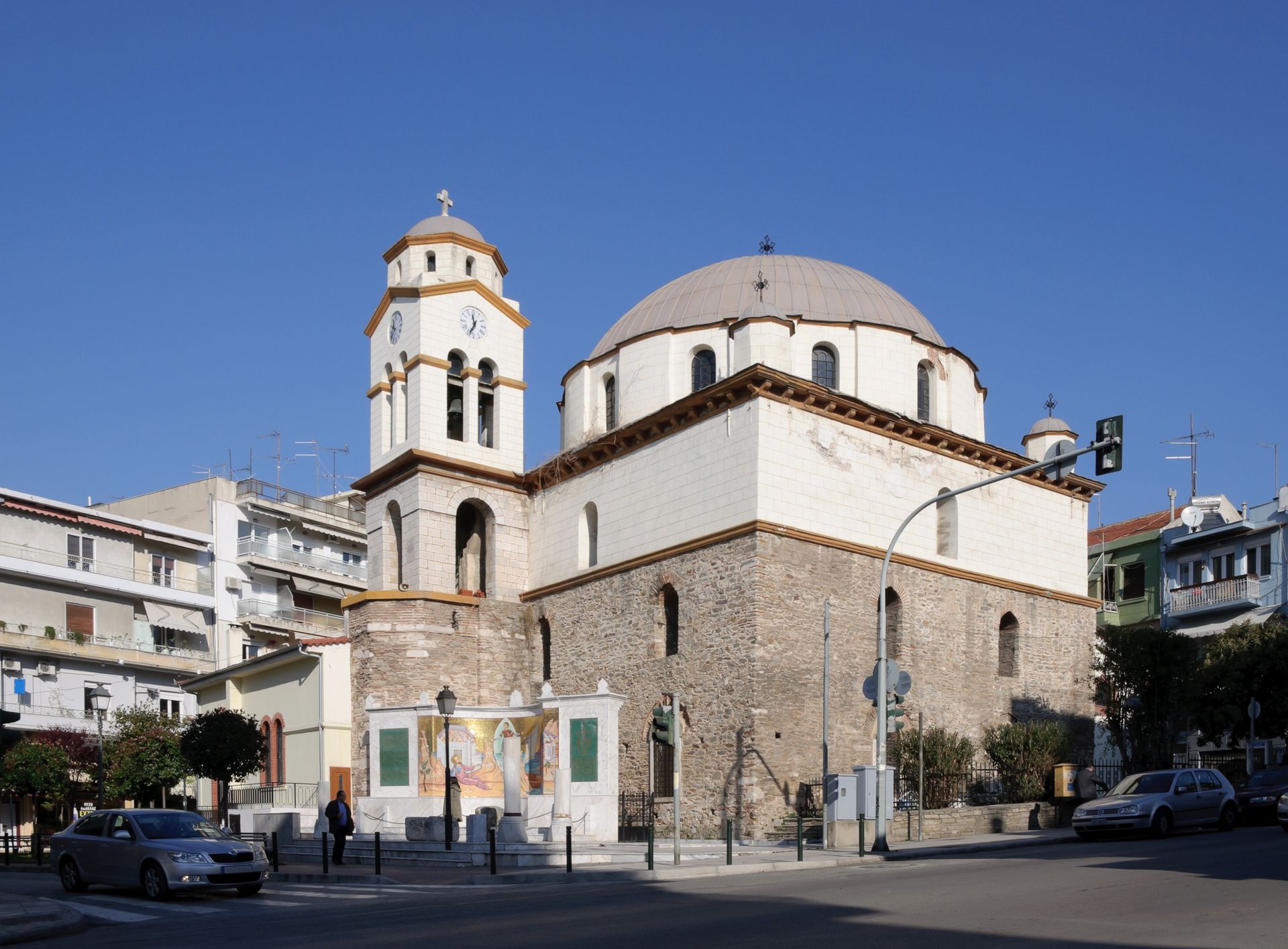 Μια ιστορική εκκλησία με πέτρινους τοίχους, μεγάλο κεντρικό τρούλο και ψηλό καμπαναριό κάτω από έναν καταγάλανο ουρανό σε ένα αστικό περιβάλλον.