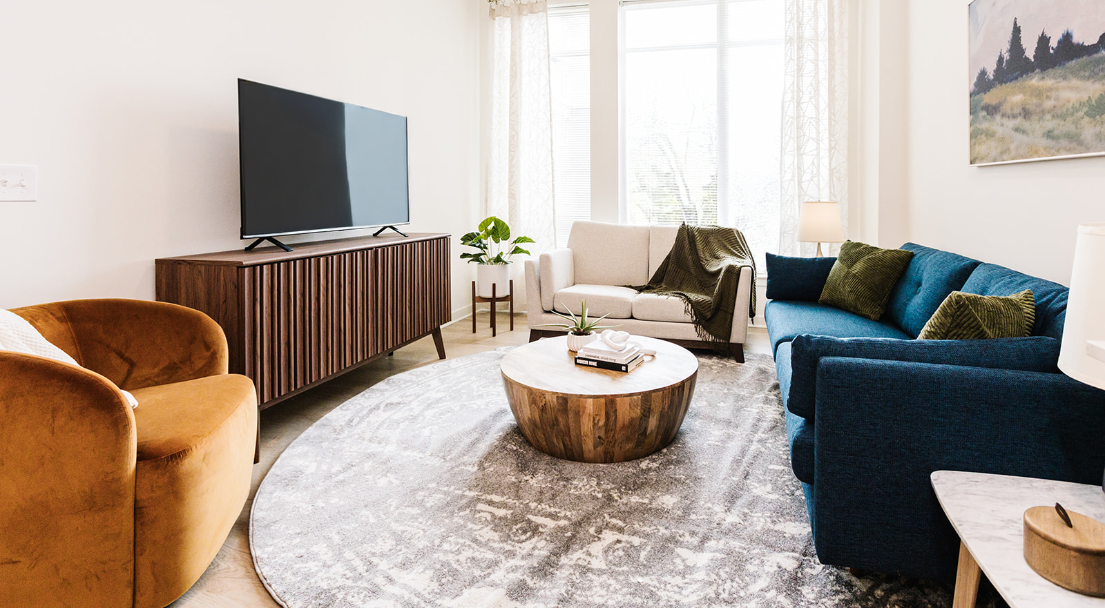 Bright living room with teal sofa, mustard chair, wooden TV stand, and round coffee table on a rug at Opal Largo Station Apartments in Largo, MD