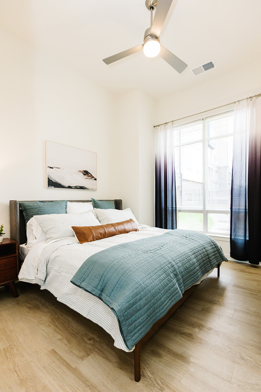 Bright bedroom with teal bedding, white walls, wood floor, and a window with sheer curtains and dark drapes at Opal Largo Station Apartments in Largo, MD