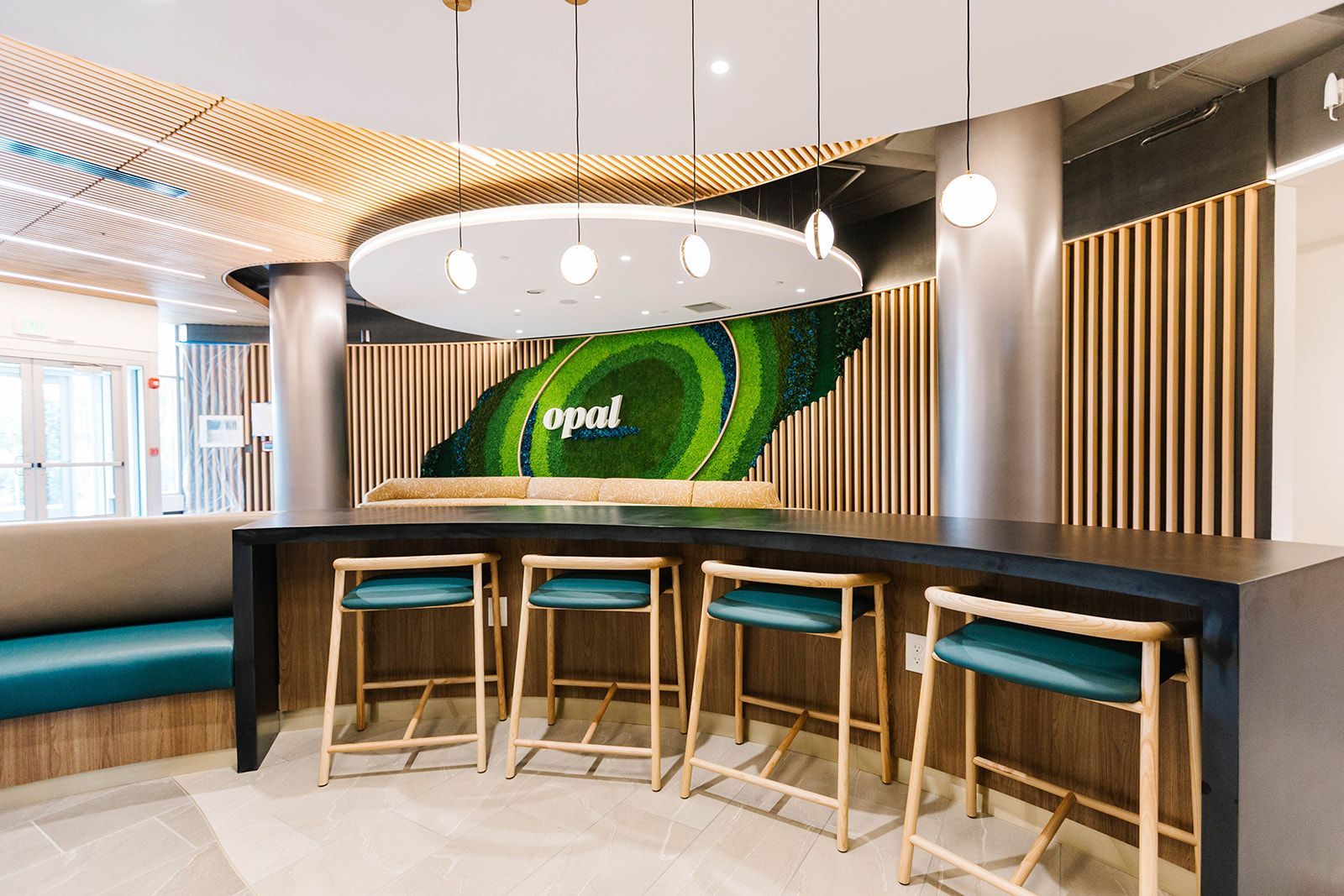 Modern cafe interior with curved wooden bar, teal stools, hanging lights, and green wall art at Opal Largo Station Apartments in Largo, MD
