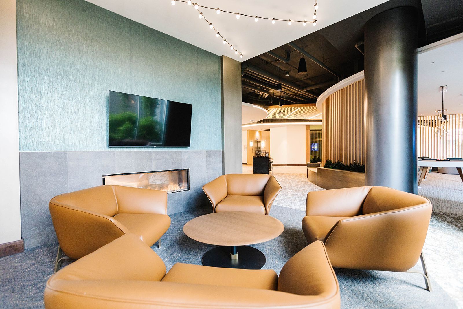 Modern lounge with tan armchairs, round tables, wall-mounted TV, and a black column in a bright interior at Opal Largo Station Apartments in Largo, MD