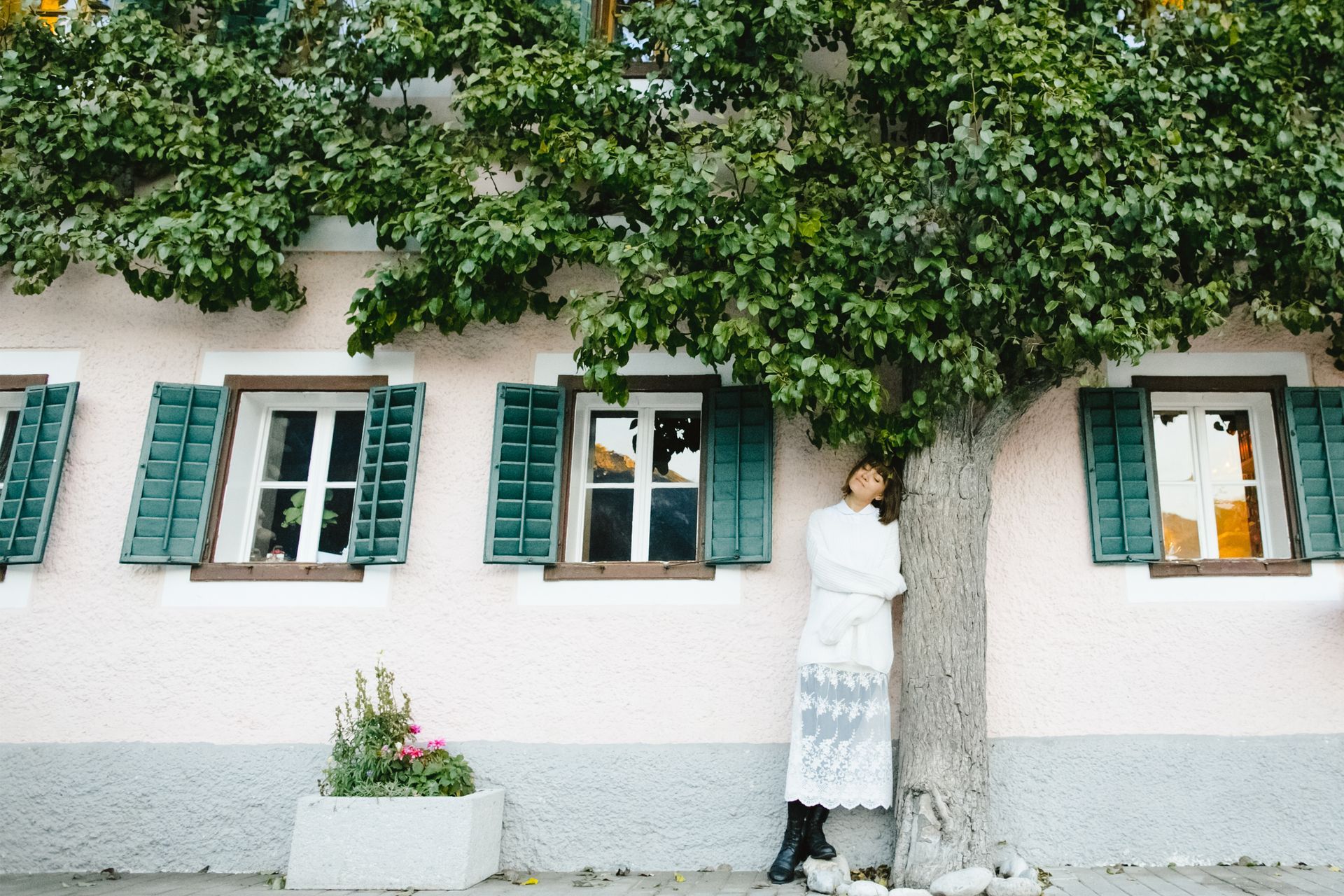 A woman leaning against a tree in front of a building with green shutters