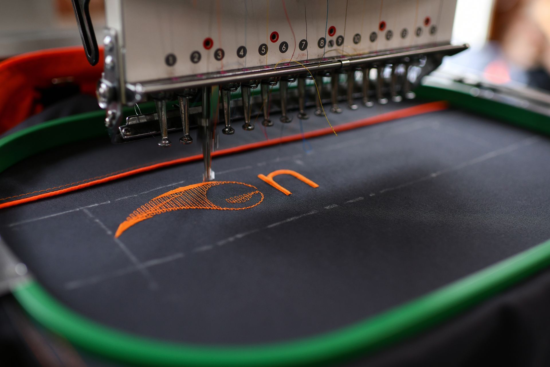 Embroidery machine stitching orange logo on black fabric, with green hoop and silver machine head.