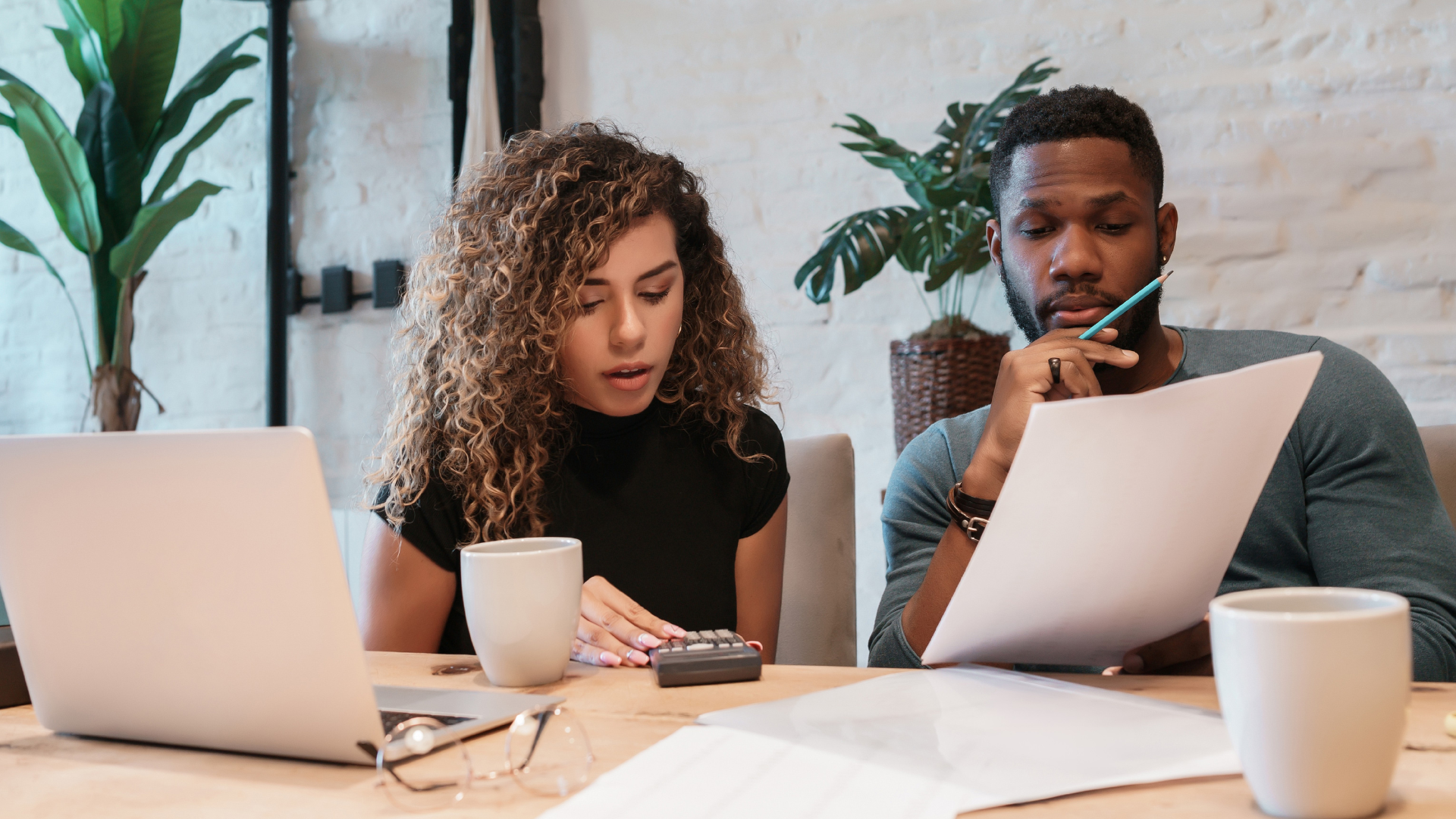 Two coworkers reviewing documents at a desk with laptops, papers, and coffee cups in a bright office.