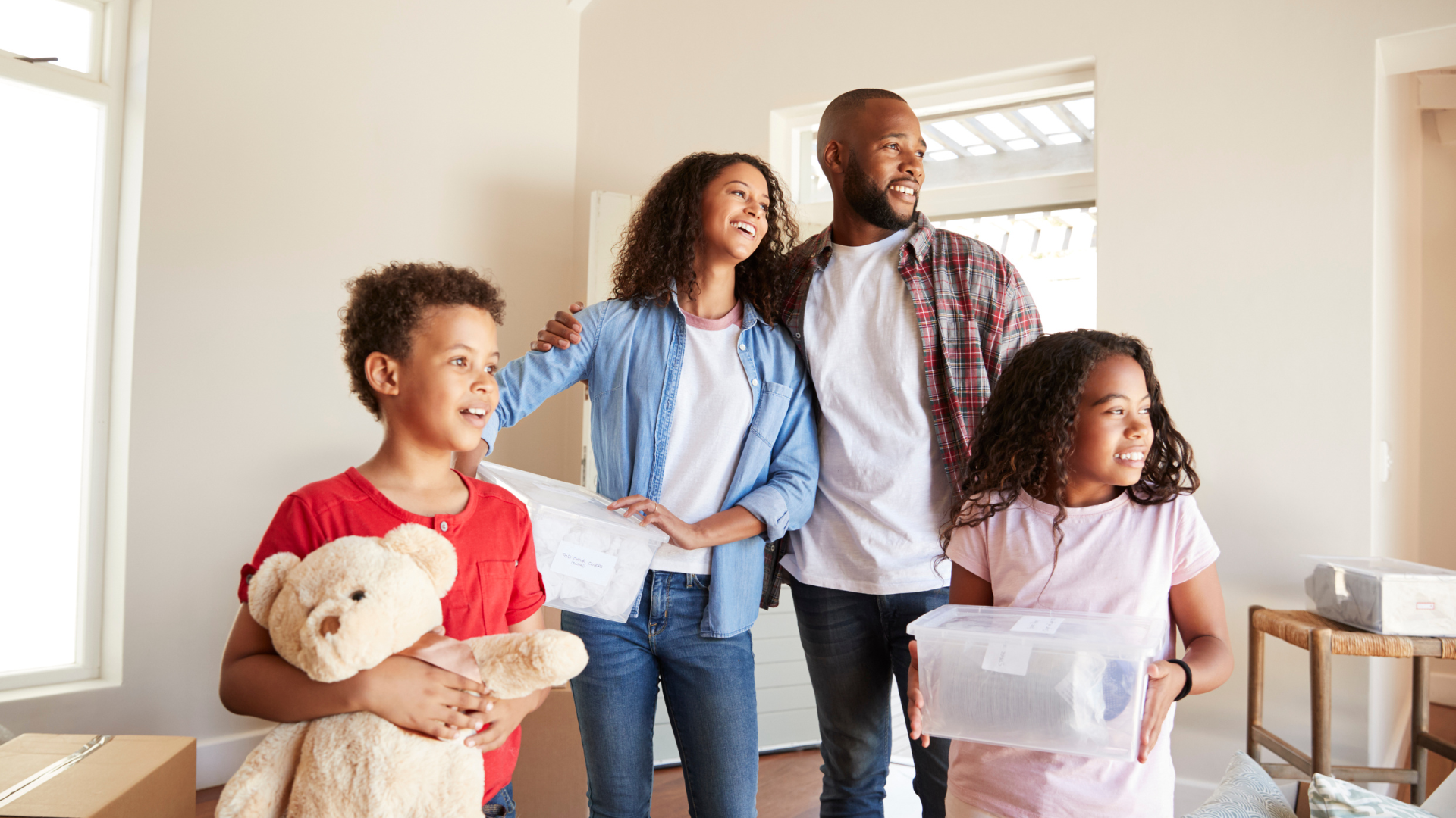 Family carrying boxes and a teddy bear in a bright room, smiling during a move.