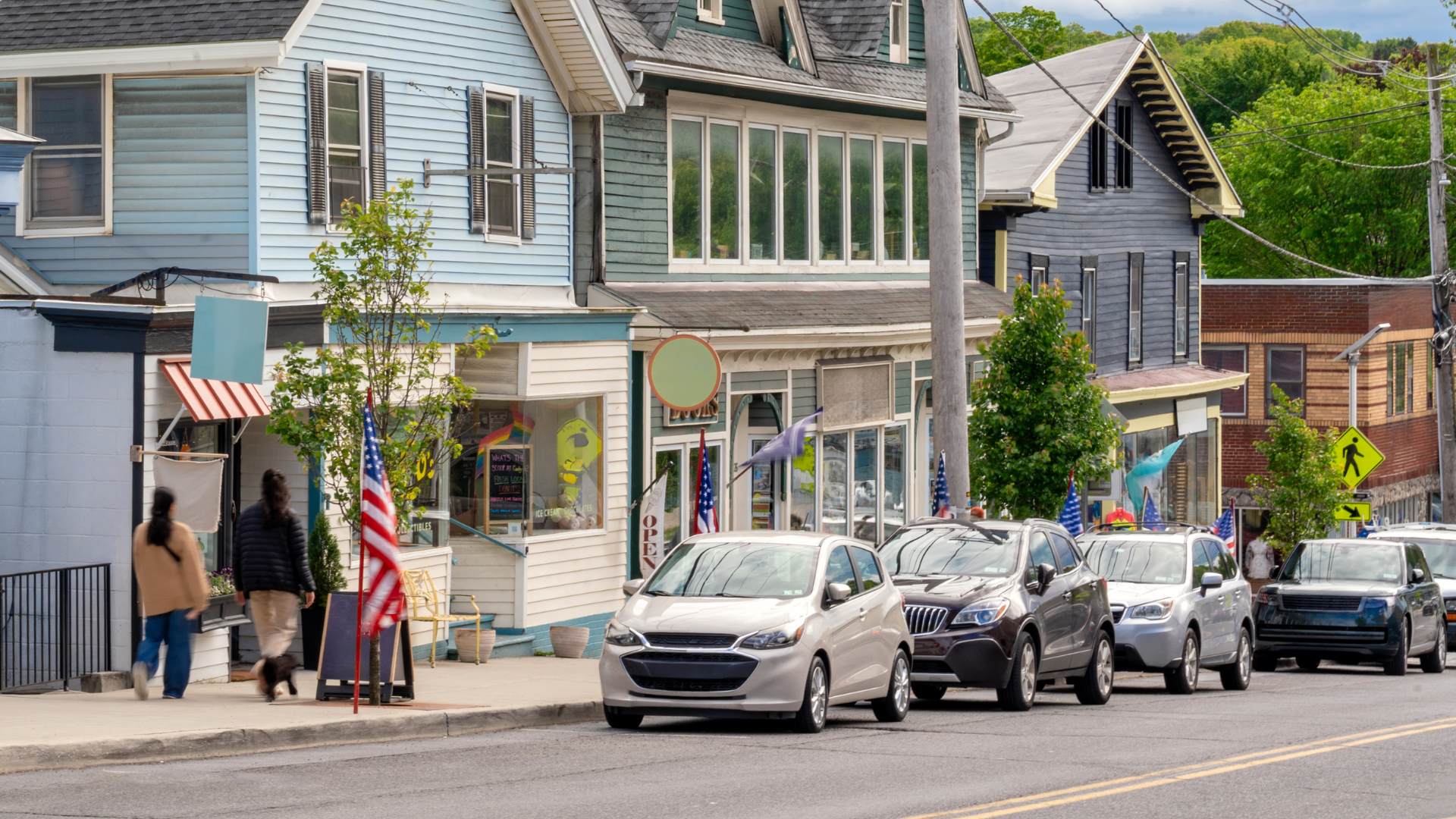 Small-town street with parked cars, pedestrians, and shopfronts under an American flag