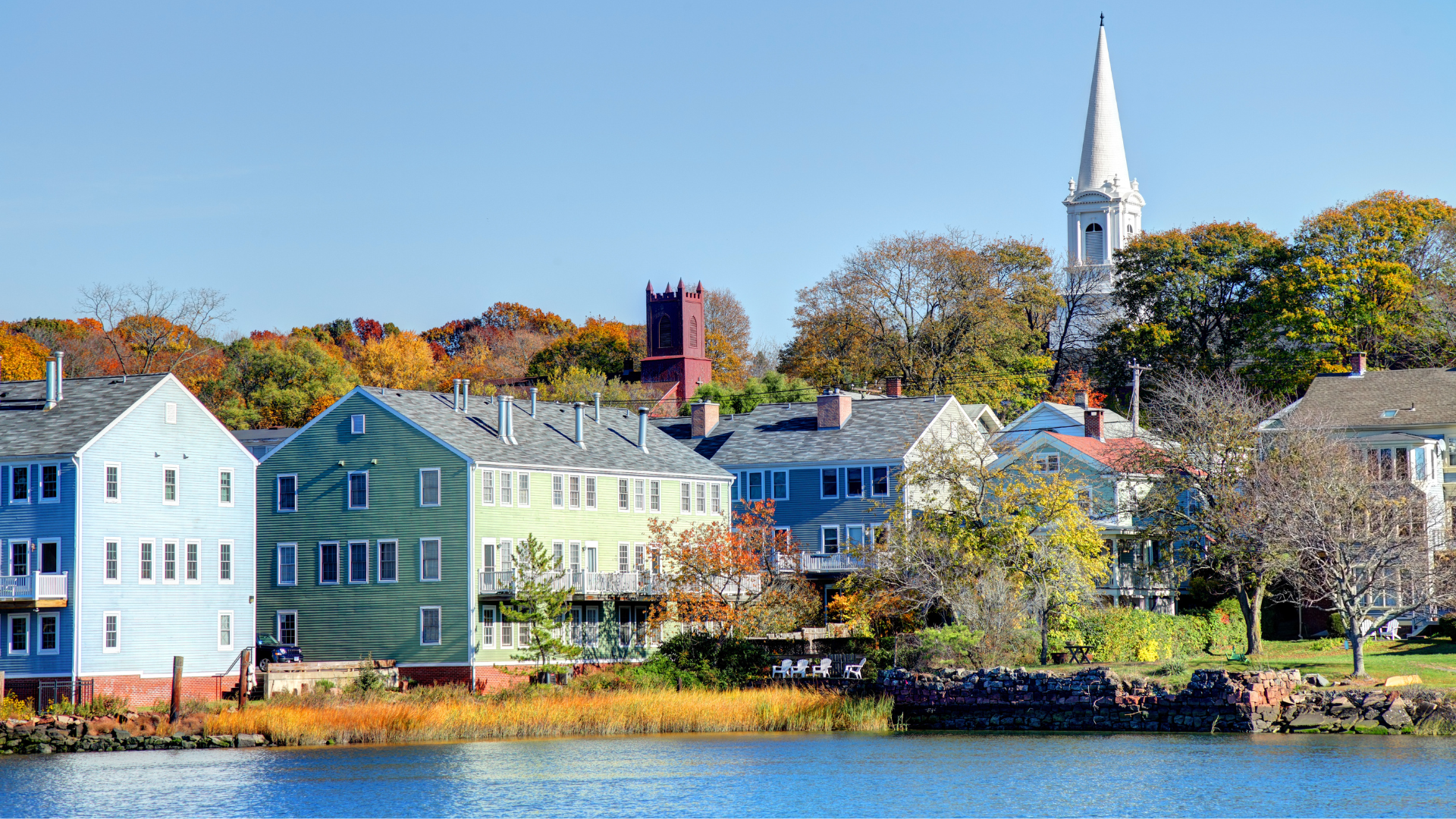 Colorful waterfront houses and a white church steeple along a calm shoreline under autumn trees