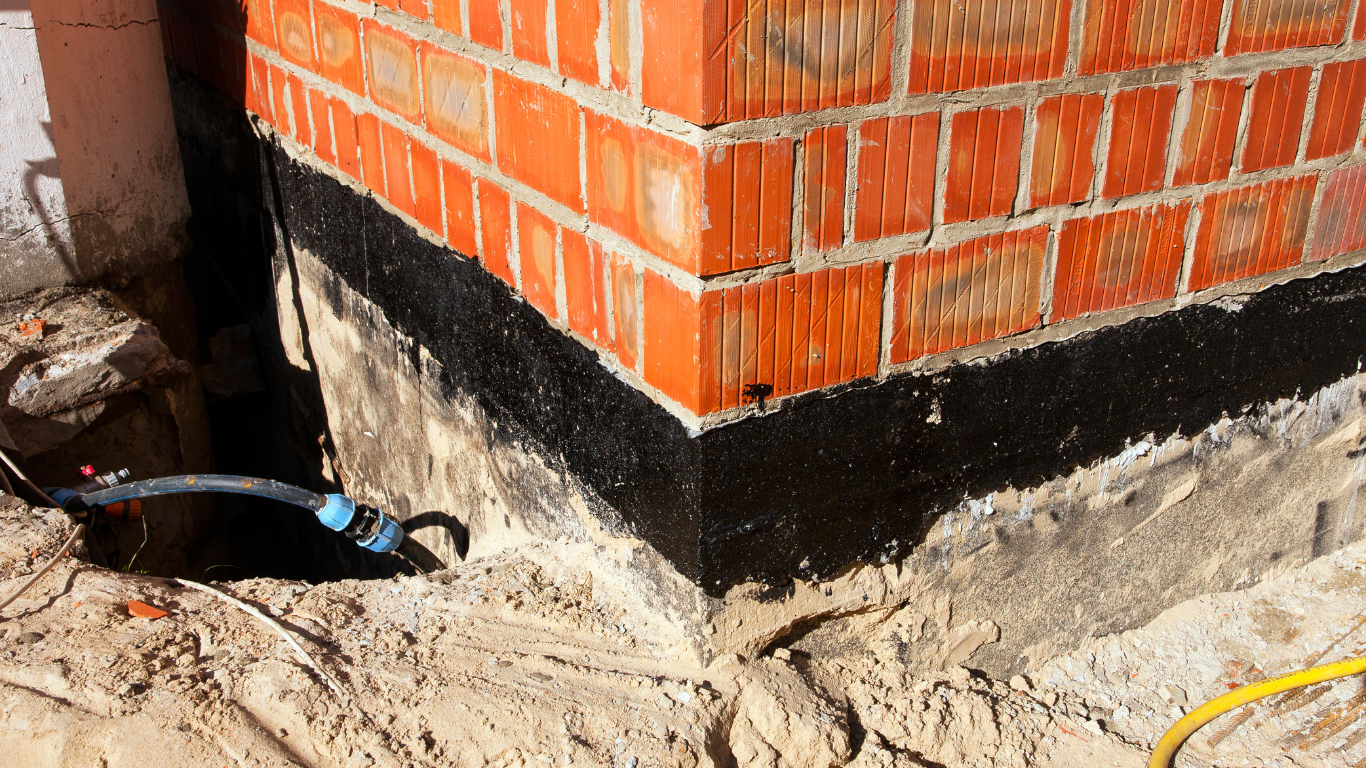 Orange brick wall with black waterproofing at the base, beside a dirt trench and exposed plumbing pipe.