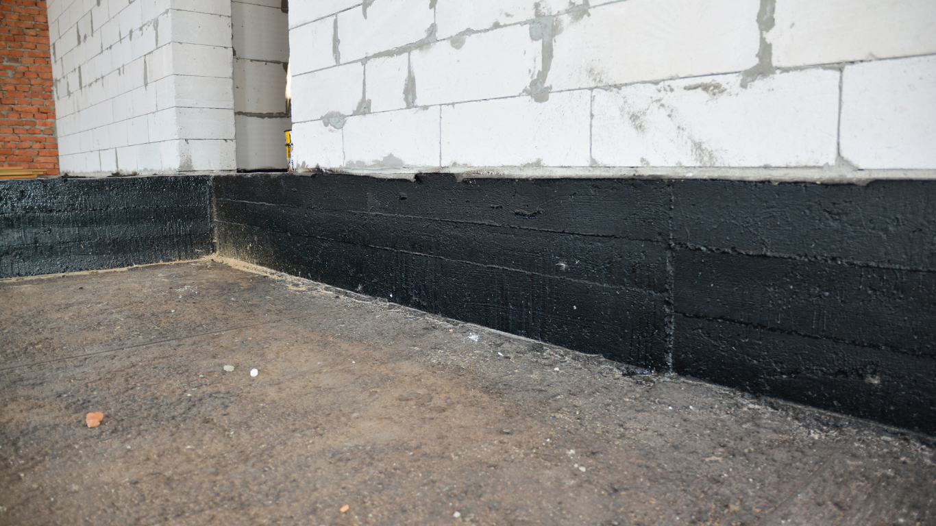Black waterproofing paint along the base of a white brick building on a concrete floor.