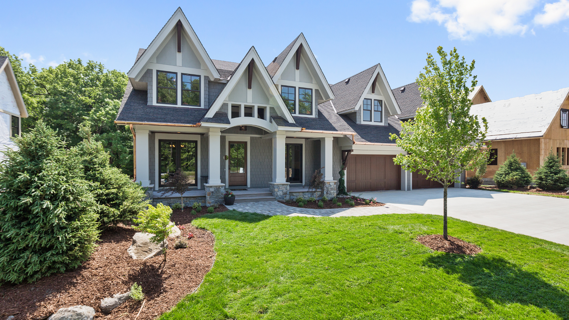Gray house with multiple gables, front porch, and garage, set on a green lawn with landscaping.