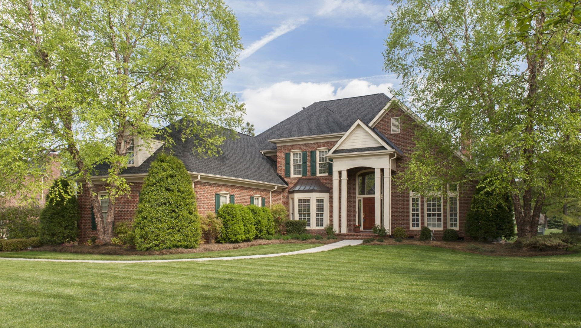 Brick suburban house with a dark roof and white columns, framed by tall trees and a green lawn