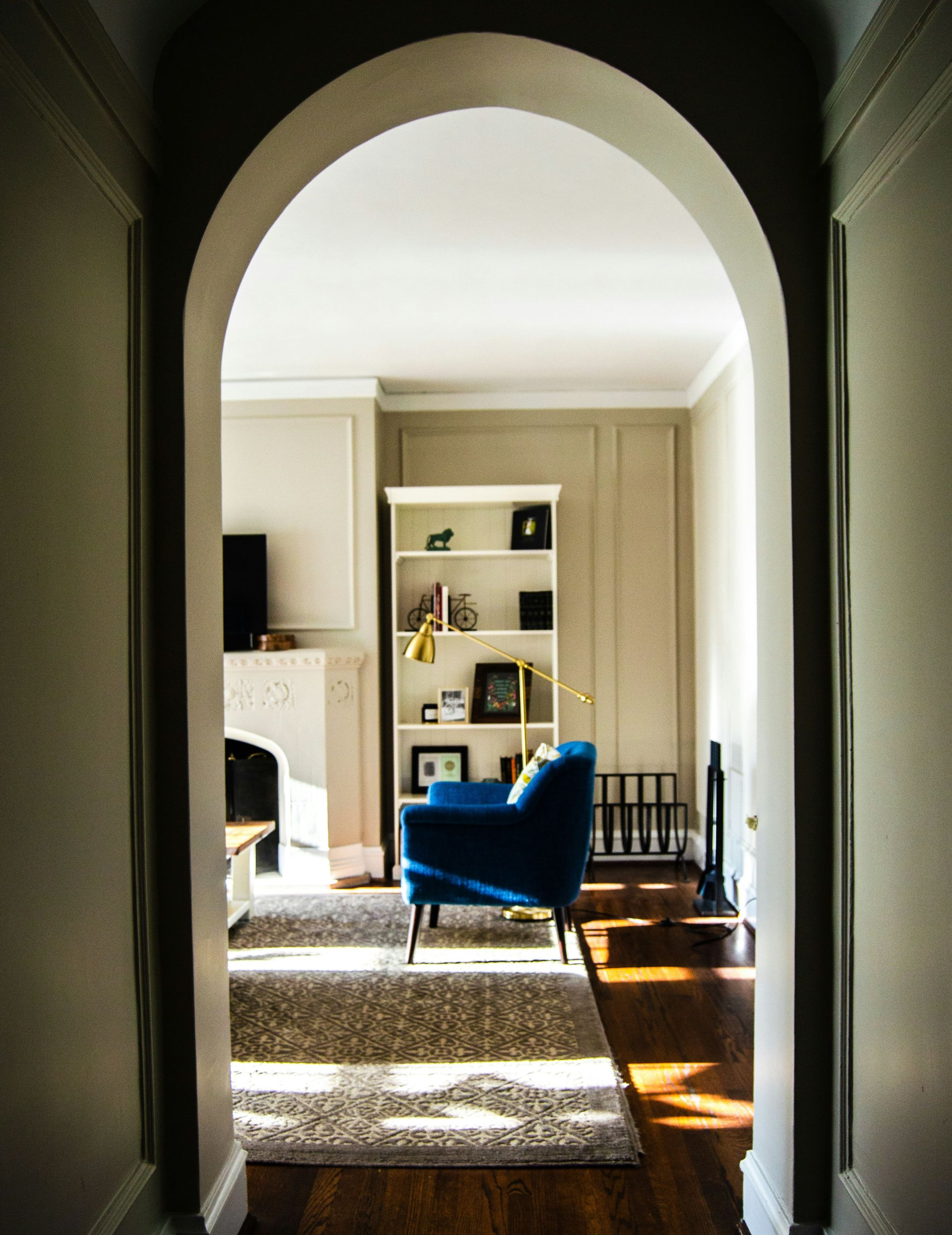 View through an arched doorway into a sunlit living room featuring a blue armchair, white bookshelf, and patterned rug.