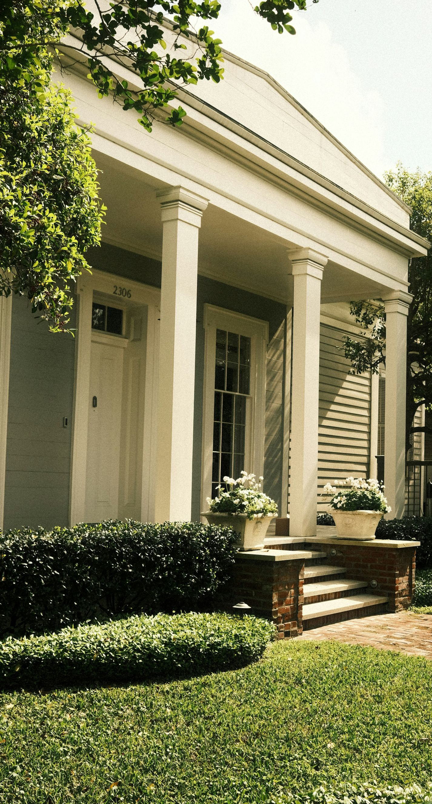 White-columned house entrance with steps, green hedges, and sunlight filtering through trees