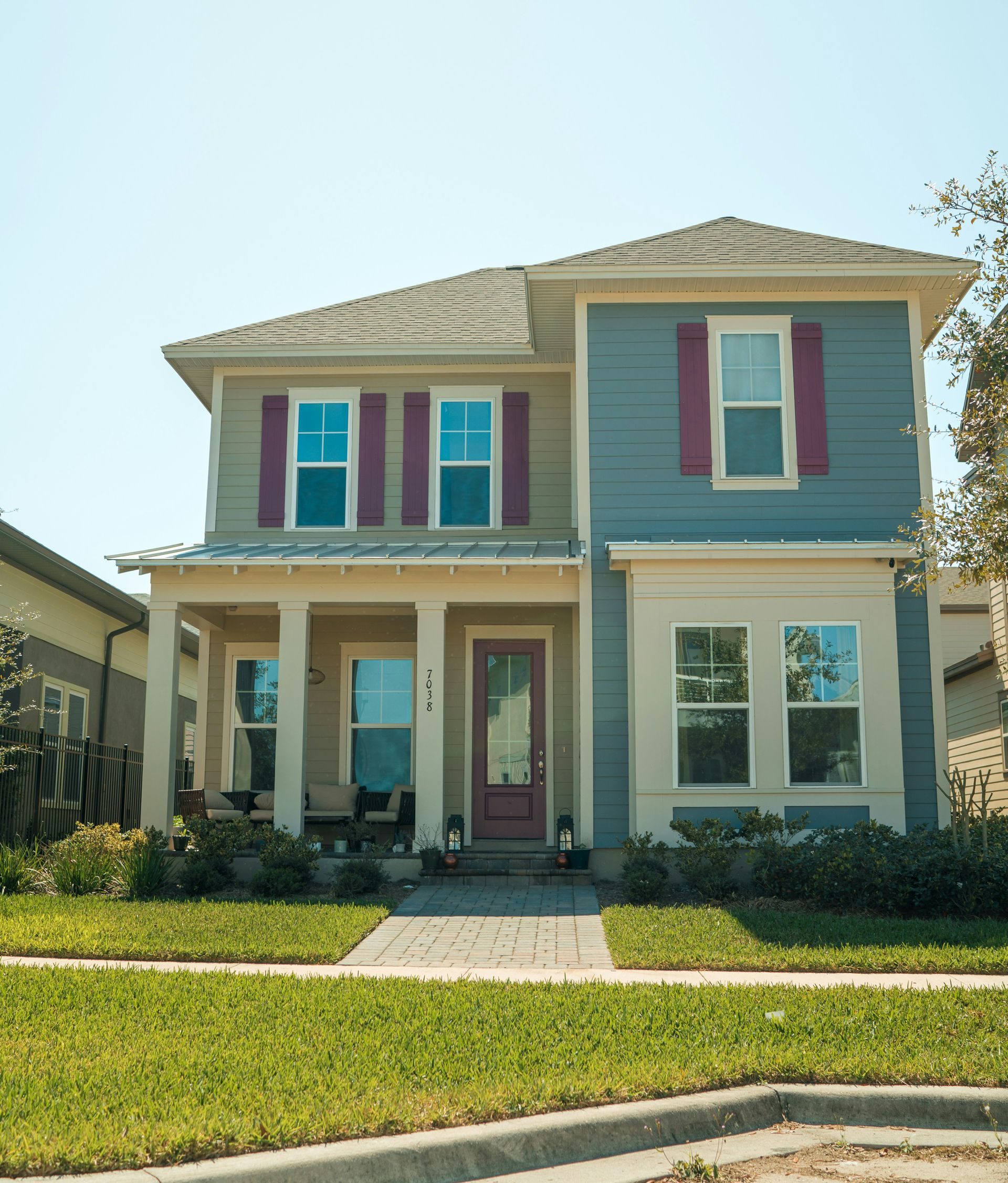 A two-story suburban house with a blue and beige facade, maroon shutters, a front porch, and a landscaped front lawn.