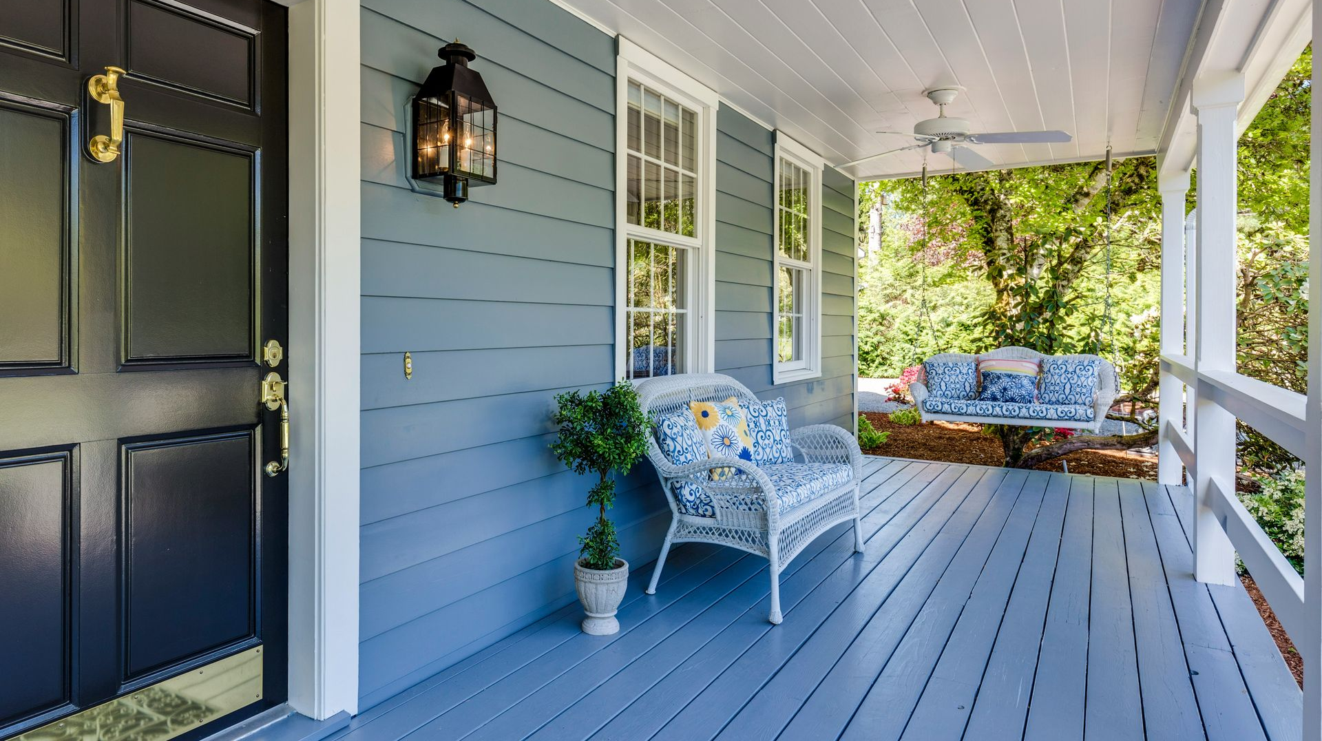 A blue-gray front porch featuring a dark blue door, white wicker furniture, a hanging swing, and a black wall lantern.