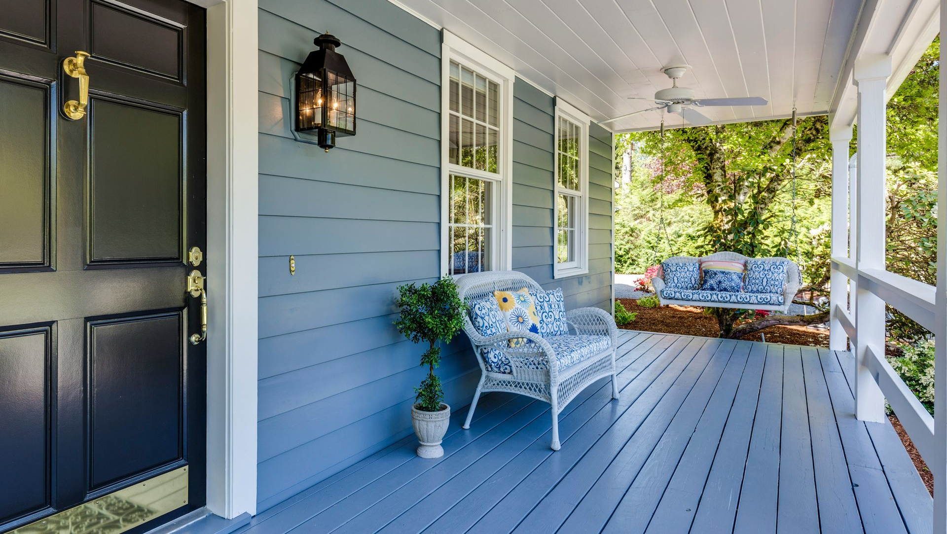 Front porch with gray siding, black door, white wicker chair, blue rug, and potted plant in bright daylight