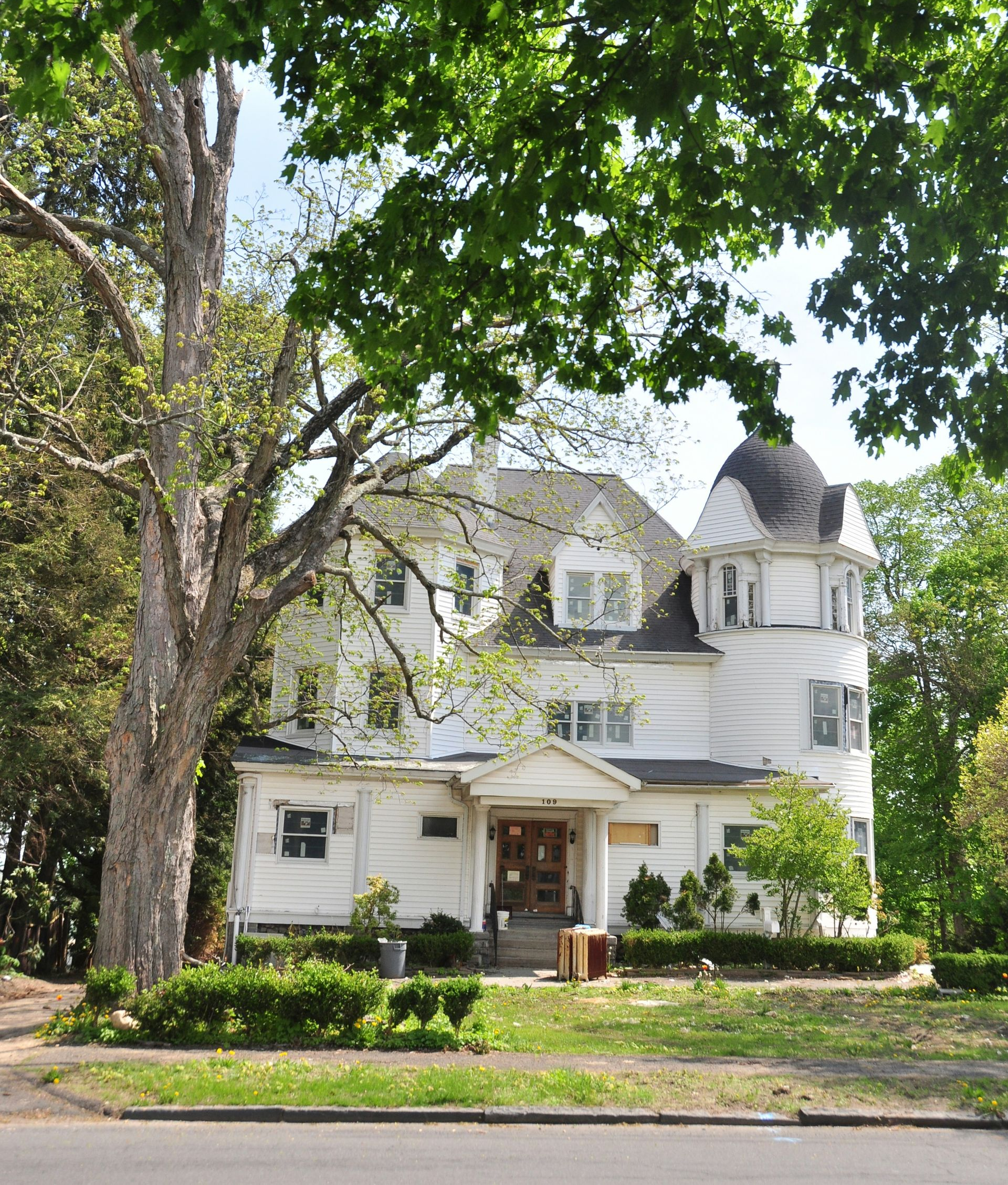 White historic house with porch and turret behind large trees on a leafy street