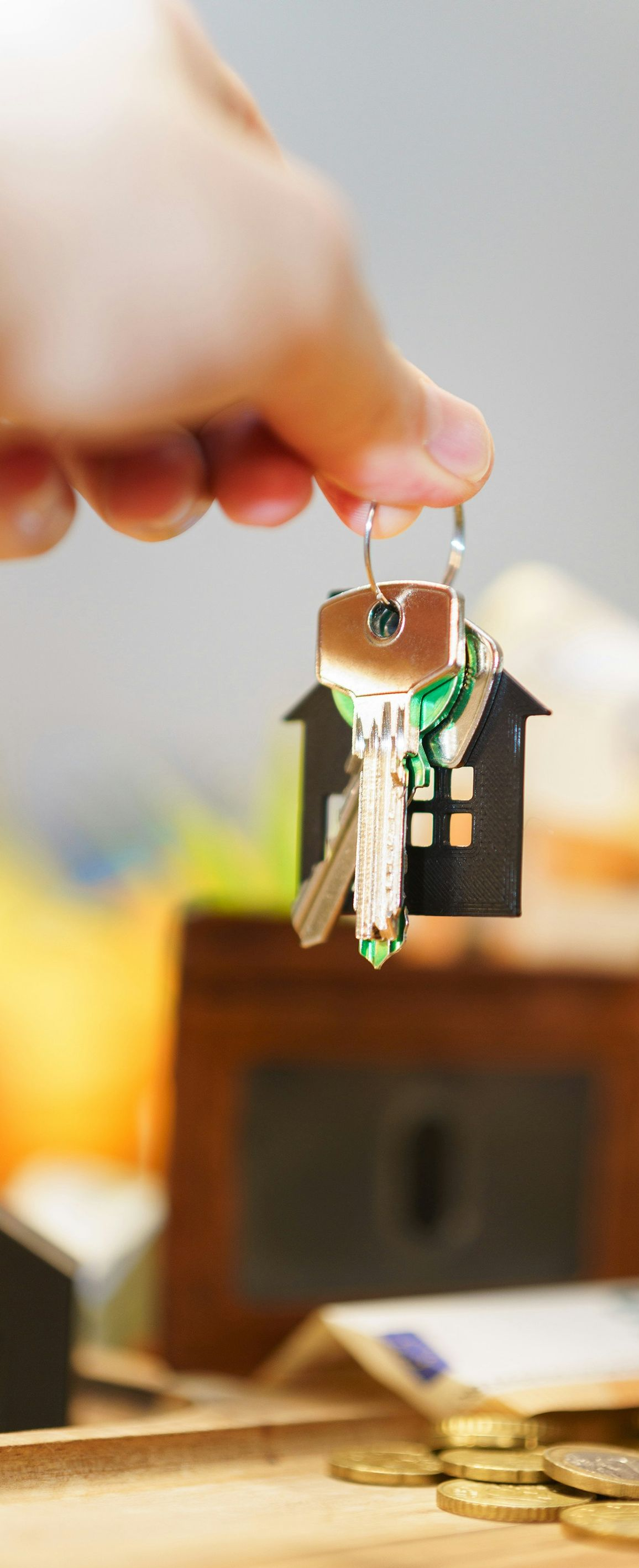 A hand holds a silver key attached to a house-shaped keychain above a brown wallet and scattered coins.