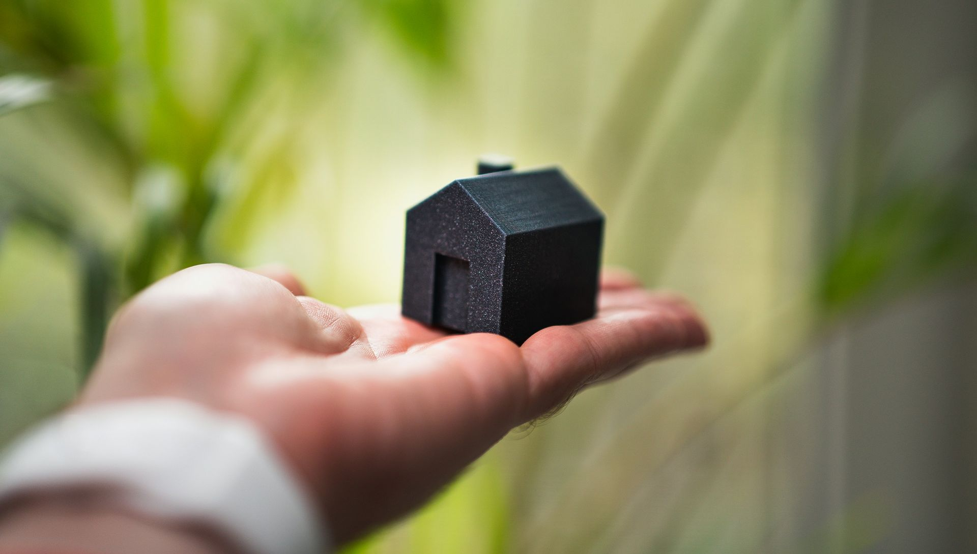 A person's hand holding a small black model house against a blurry green foliage background.