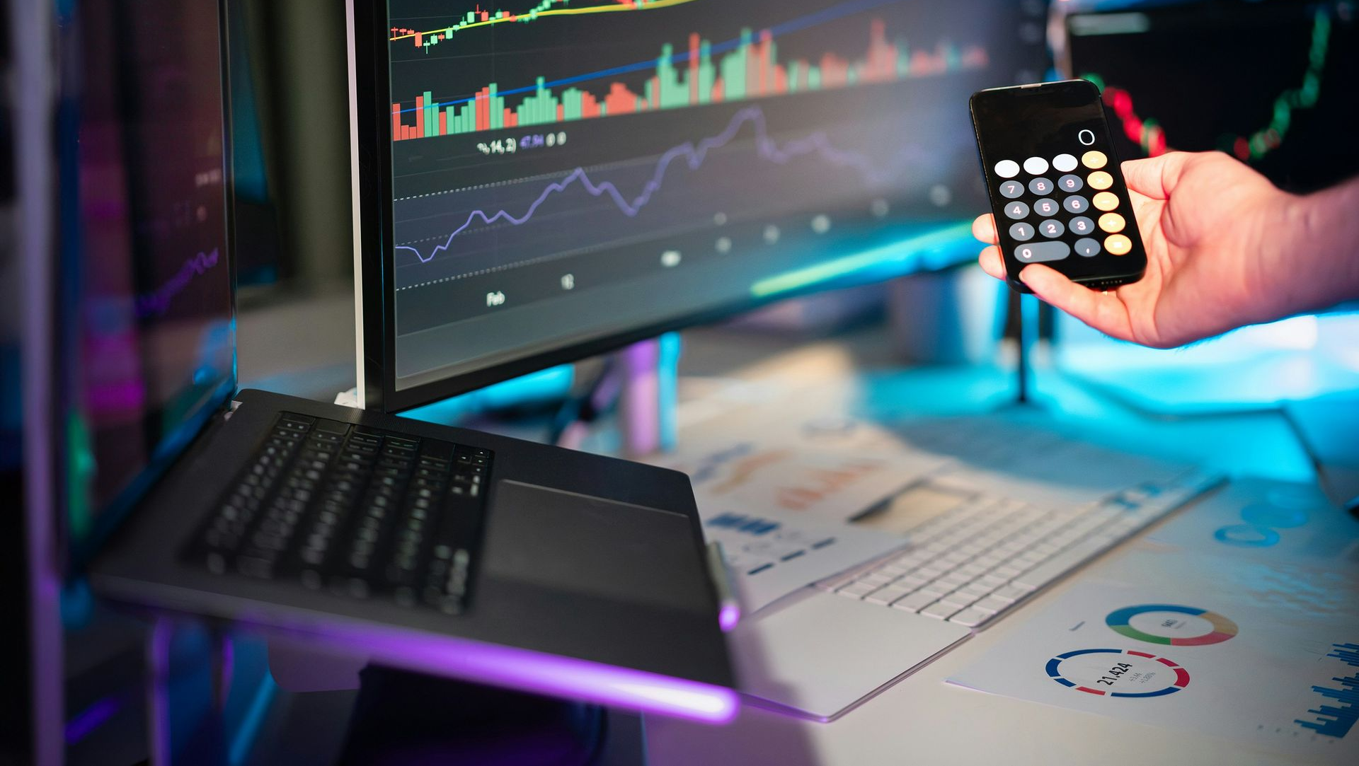 A hand holding a calculator in front of computer monitors displaying stock market charts and data in a dimly lit office.