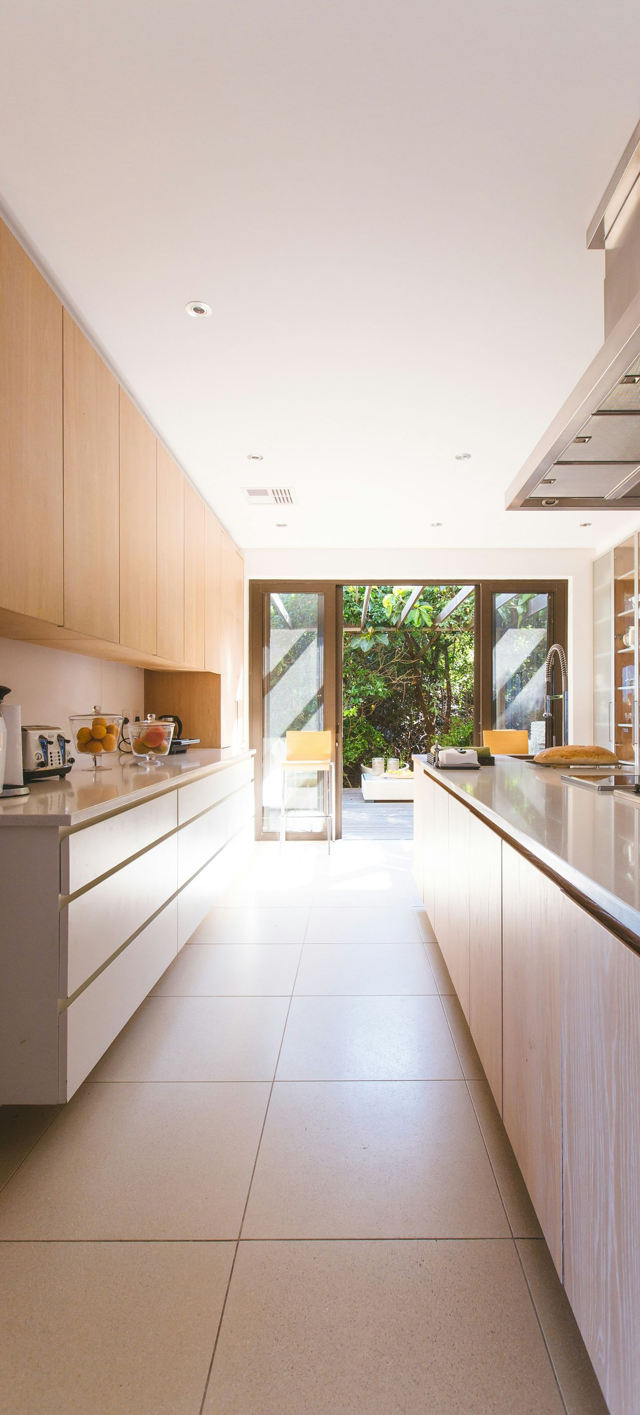 Bright modern galley kitchen with wood cabinets, long counter, and open glass doors to a garden.