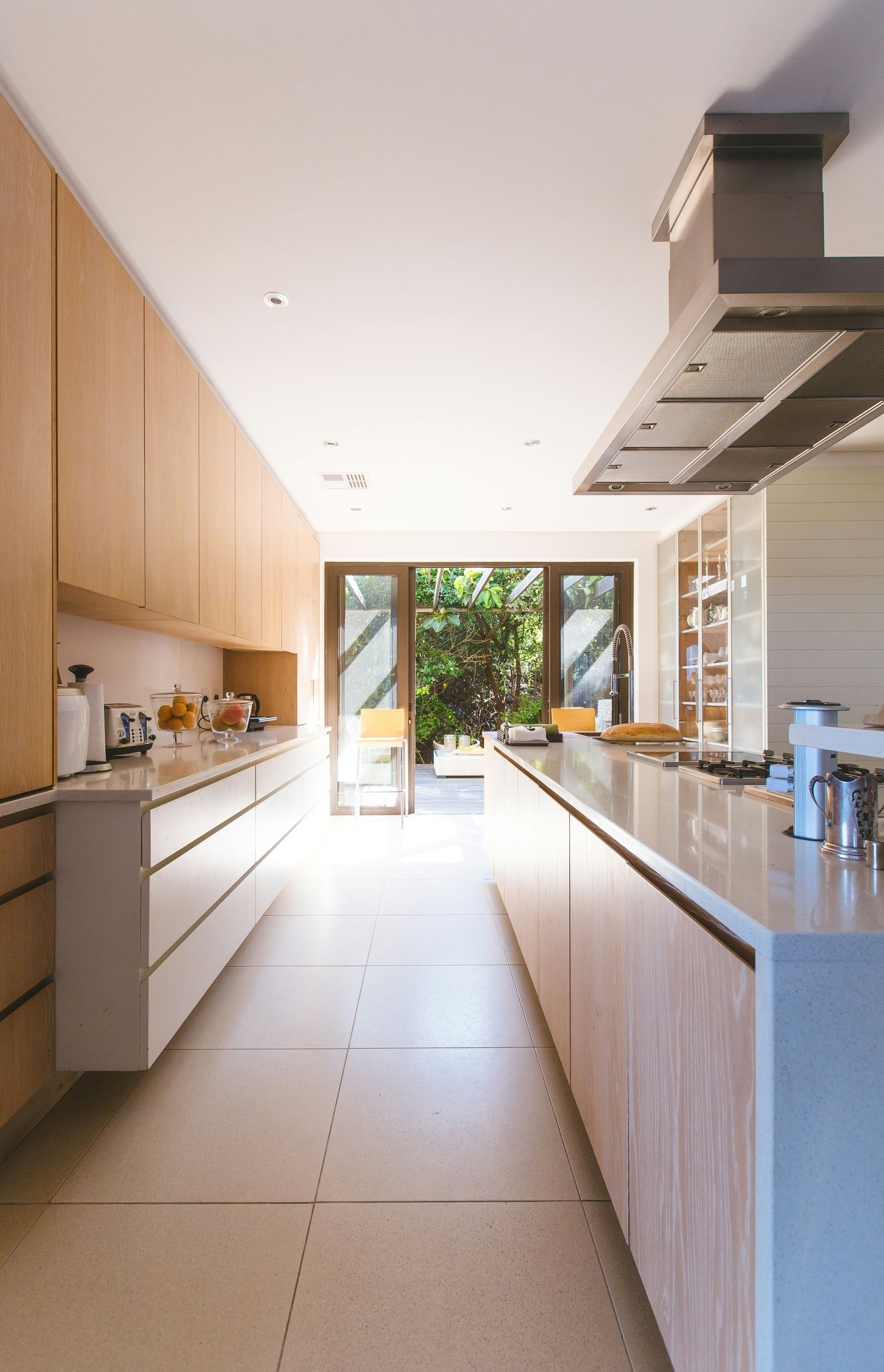 A modern galley kitchen featuring light wood cabinetry, white countertops, and a large metal range hood above an island.