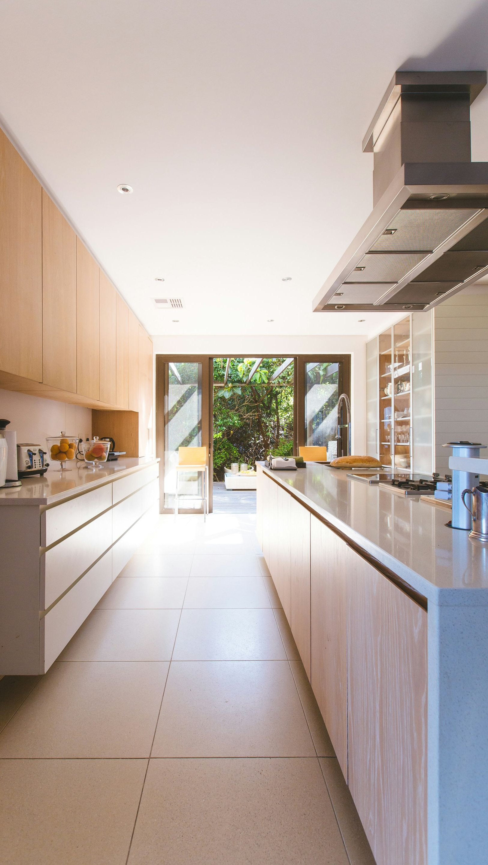 A modern galley kitchen featuring light wood cabinetry, white countertops, and a large metal range hood above an island.