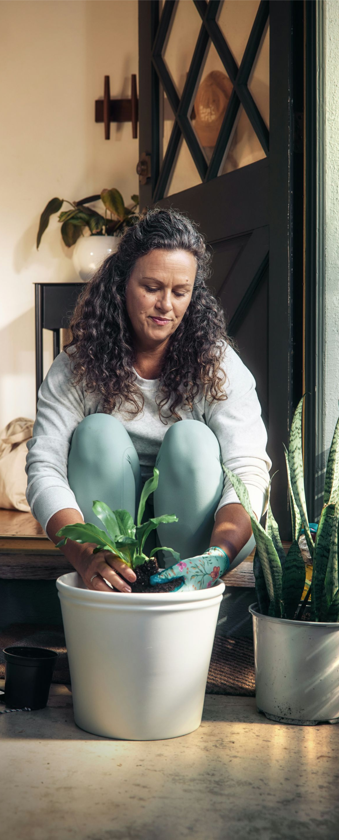 Person kneels by potted plants in a doorway, tending greenery in a sunlit indoor porch.