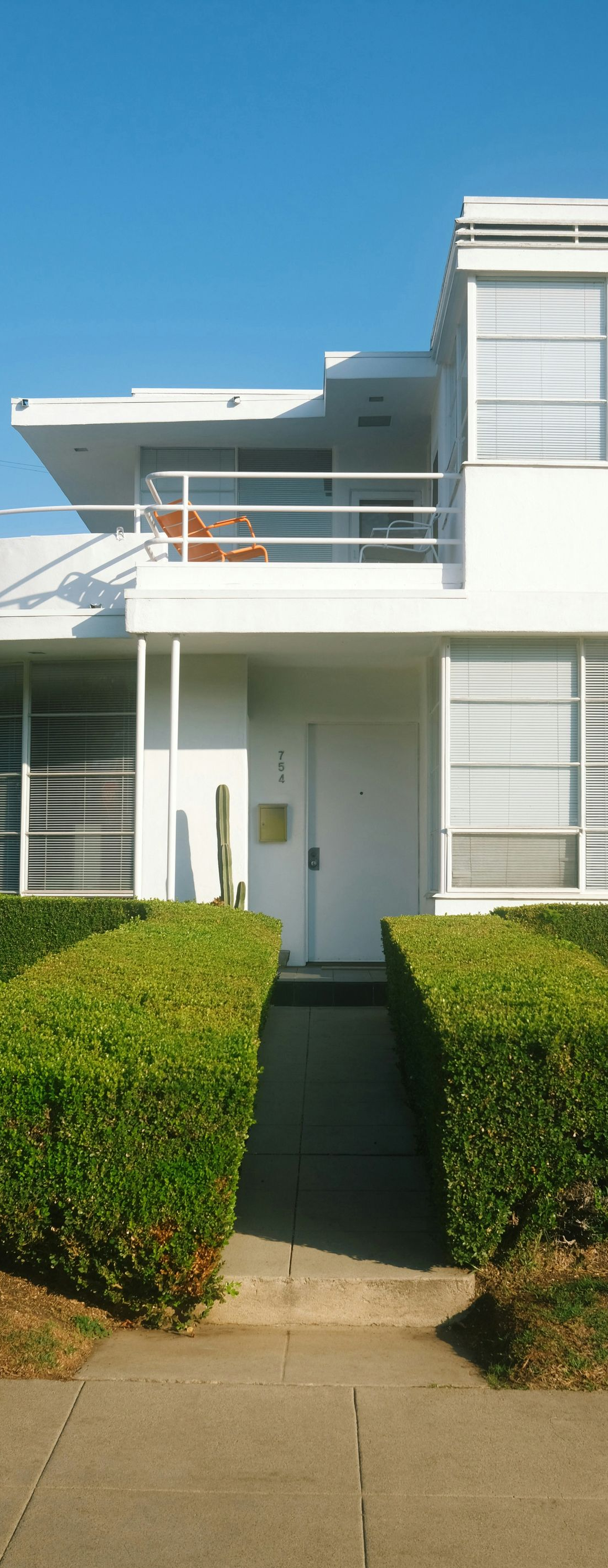 Modern white townhouse with balcony, front hedge, and blue sky