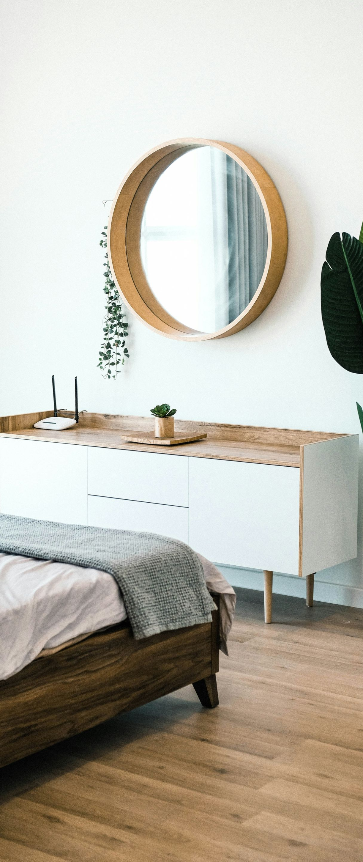 Minimal bedroom with a bed, white dresser, round mirror, and tall green plant on wood flooring