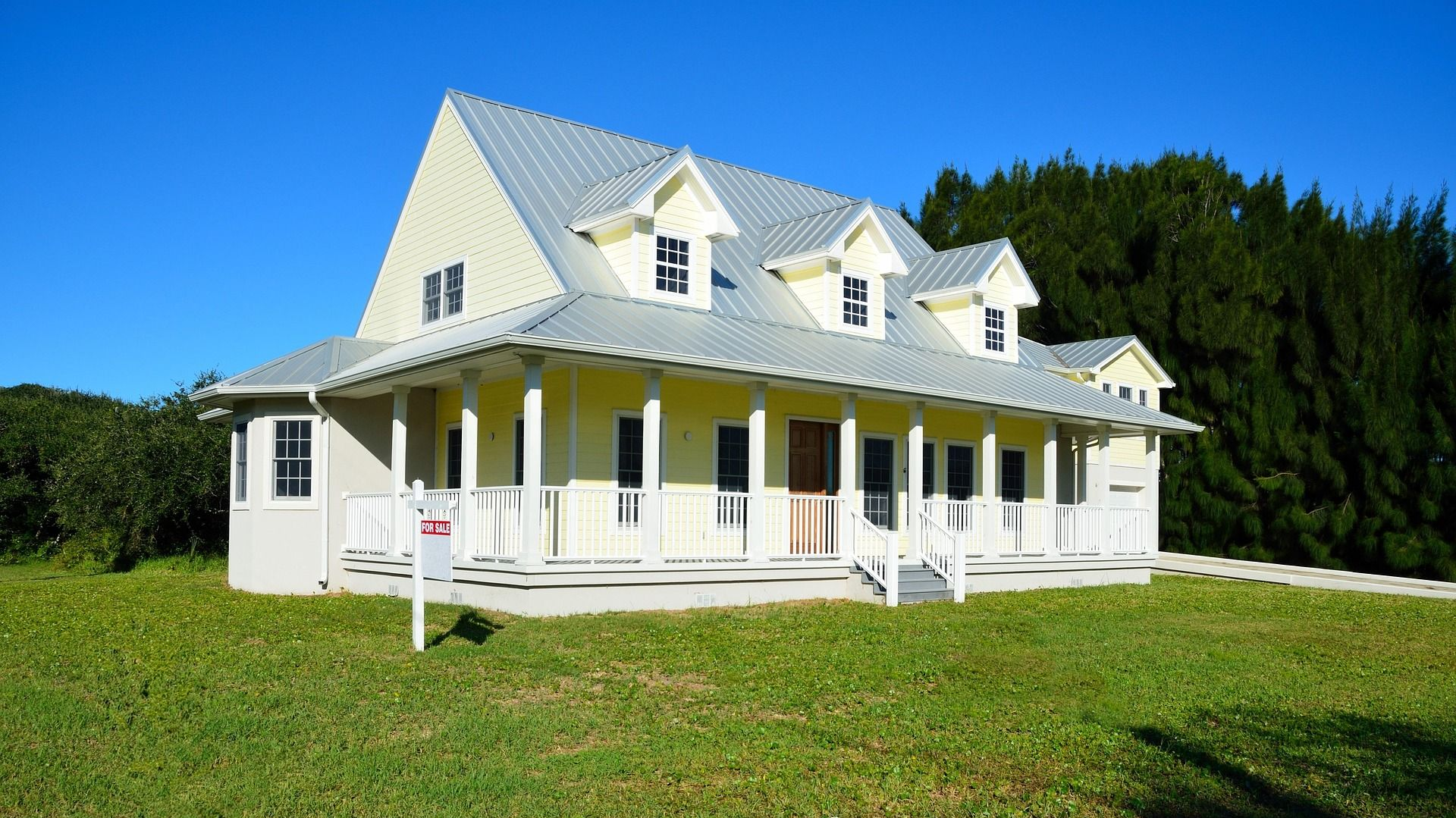 White country house with a porch and dormer windows on a grassy lawn under a blue sky