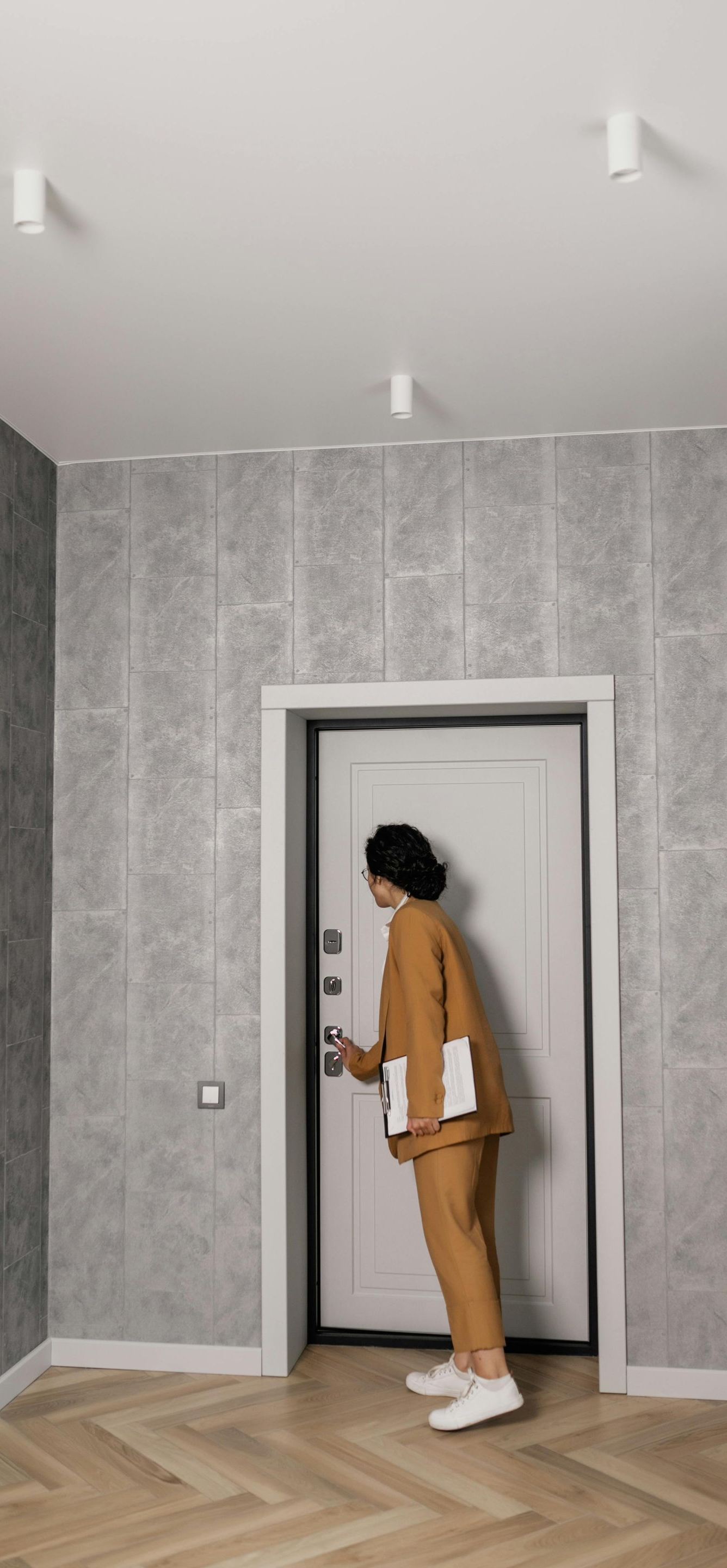 Person in a tan suit opening a white door, holding documents. Standing in a hallway with patterned gray wallpaper.