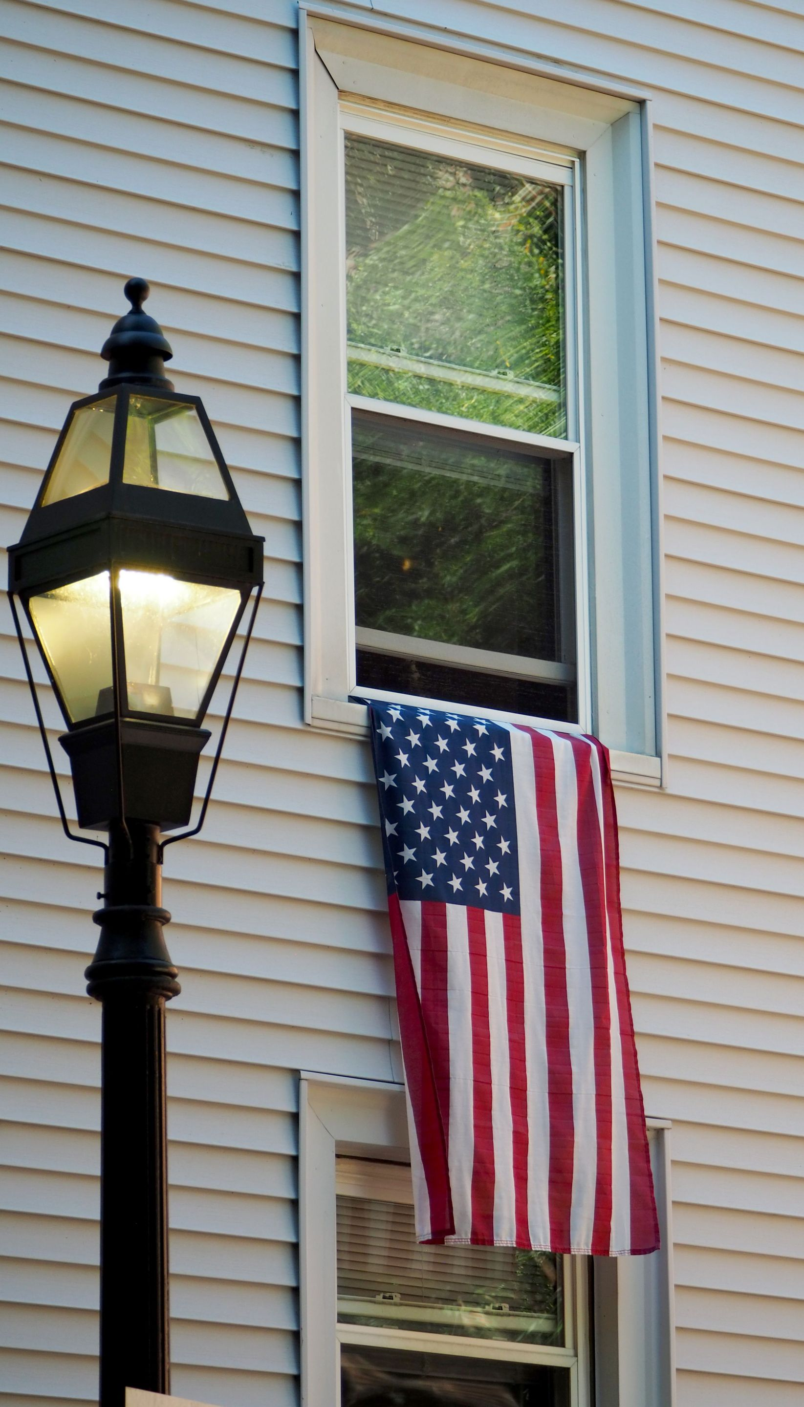 American flag hanging beside a window on a beige house, with a black lantern in the foreground