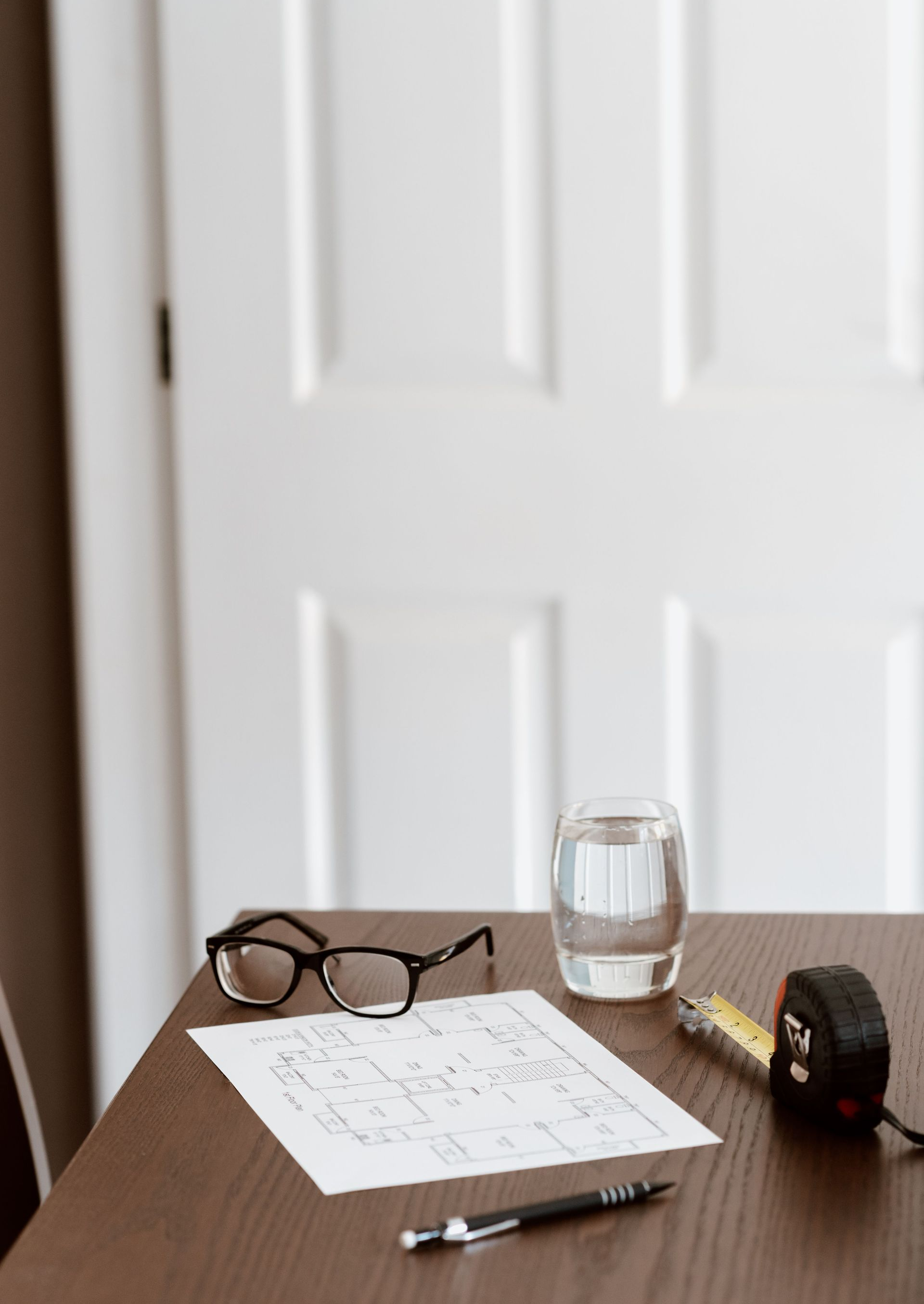 Desk with eyeglasses, papers, pen, glass of water, and tape measure in front of a white door.