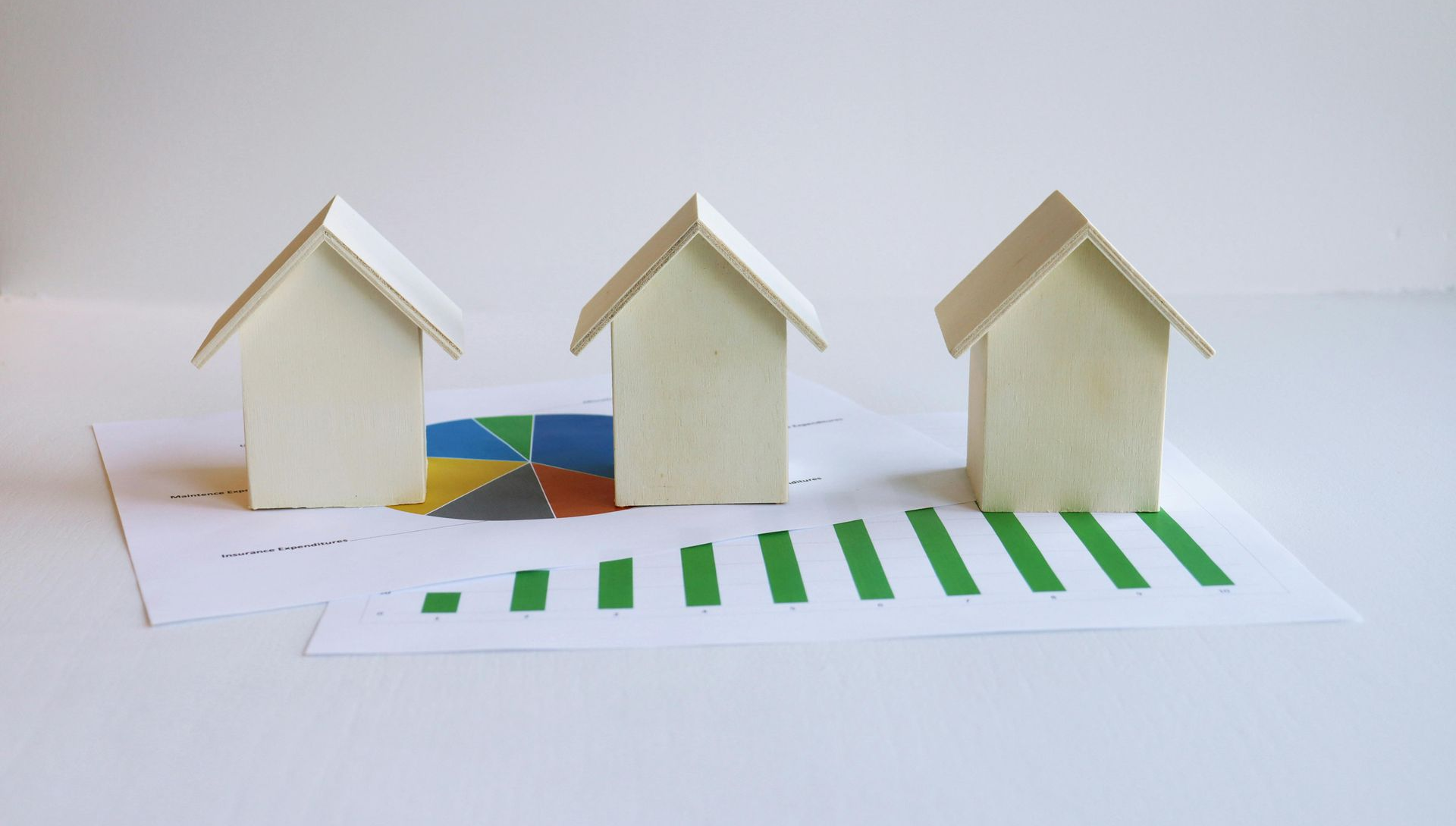 Three small wooden house models sit on a white surface atop a paper showing a colorful pie chart and a green bar graph.