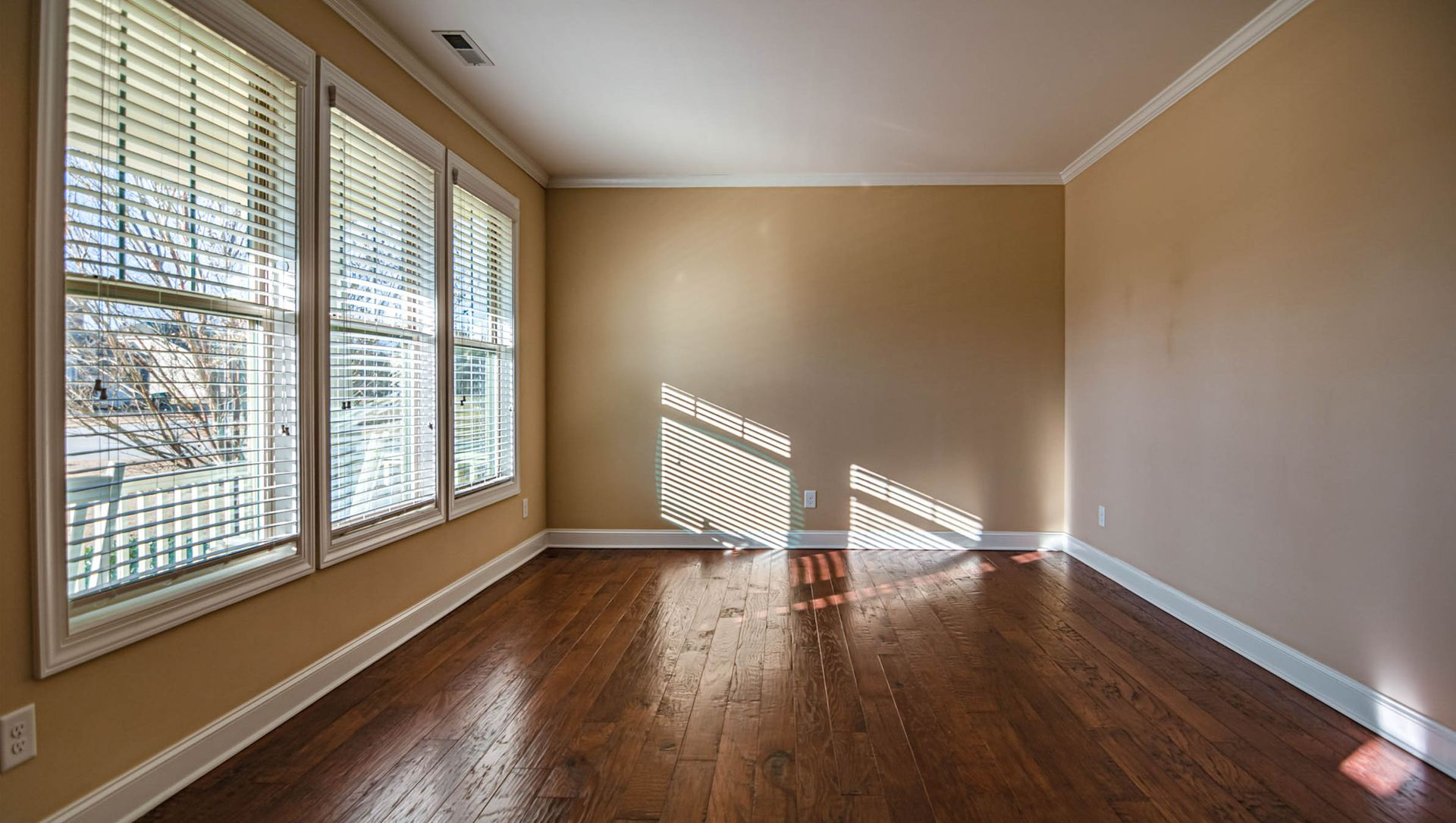 Empty beige room with hardwood floors, large windows, and sunlight casting blinds’ shadows on the wall.