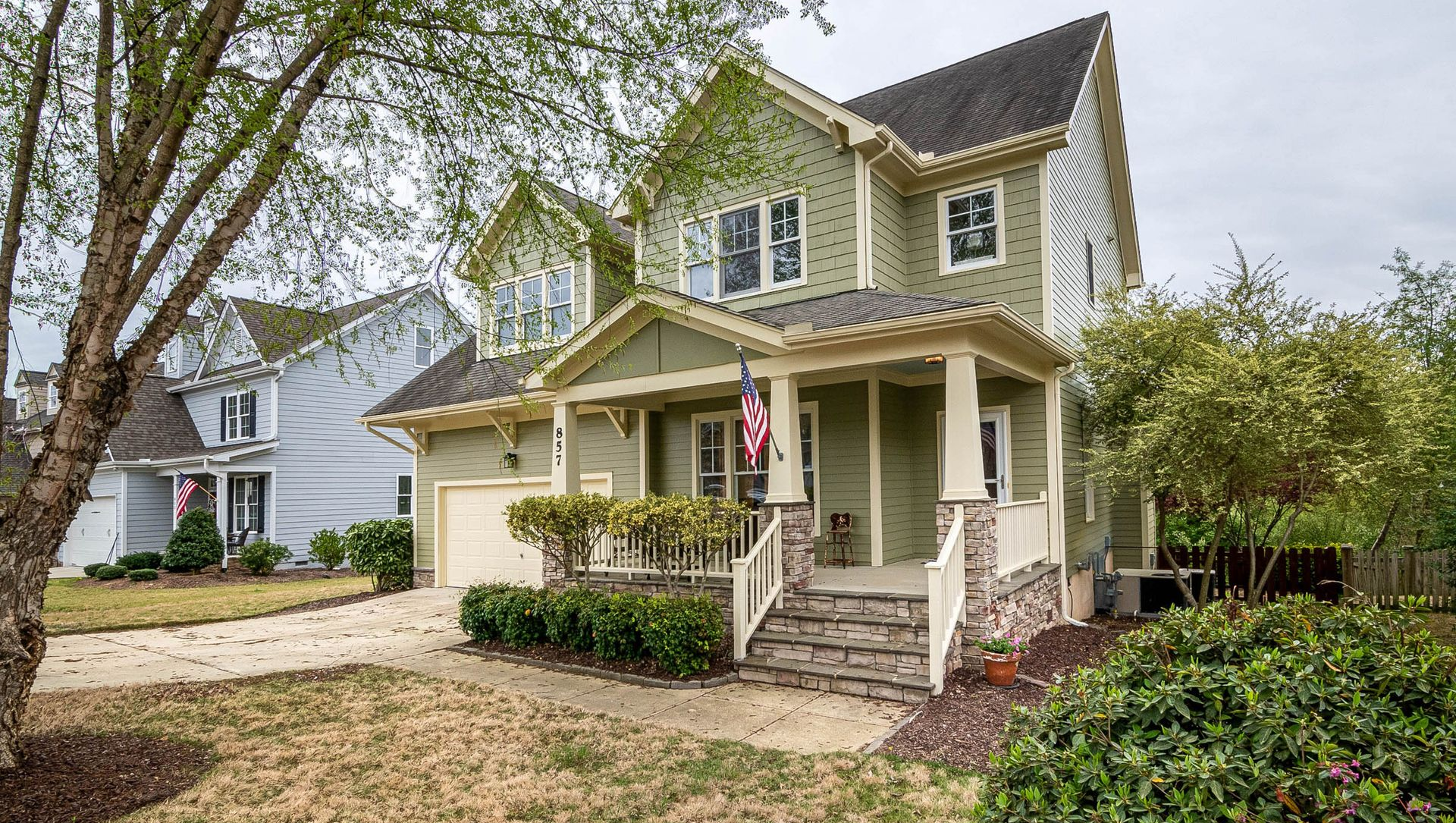 Two-story beige house with front porch, white trim, and an American flag, viewed from the street.