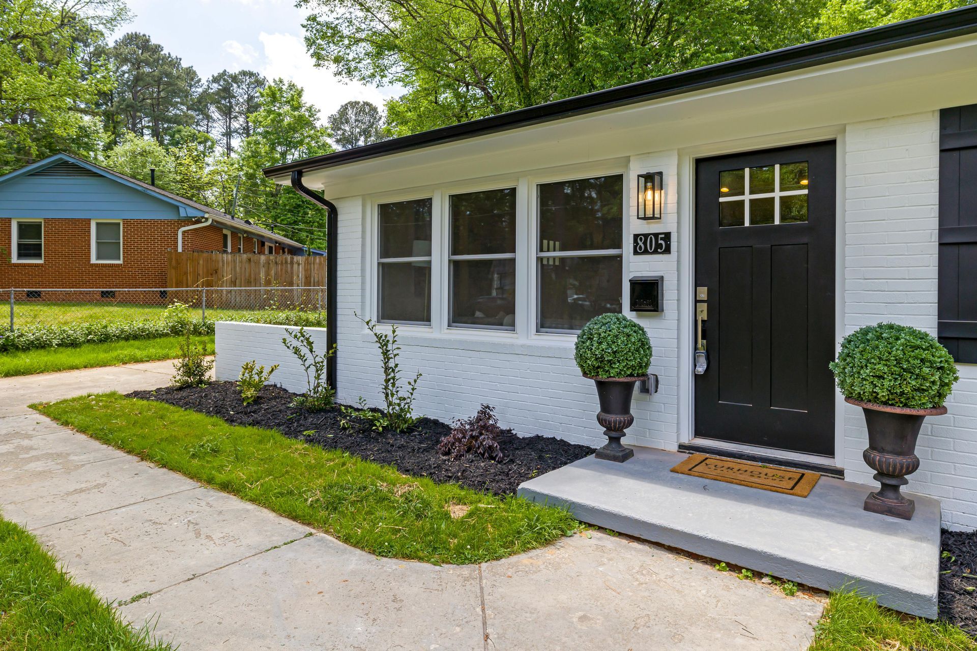 White house entrance with black door, potted plants, and a concrete walkway in a suburban neighborhood