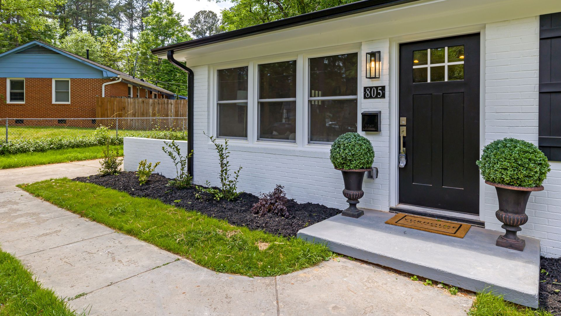 Modern white house exterior with black door, front porch, potted topiaries, and concrete walkway.