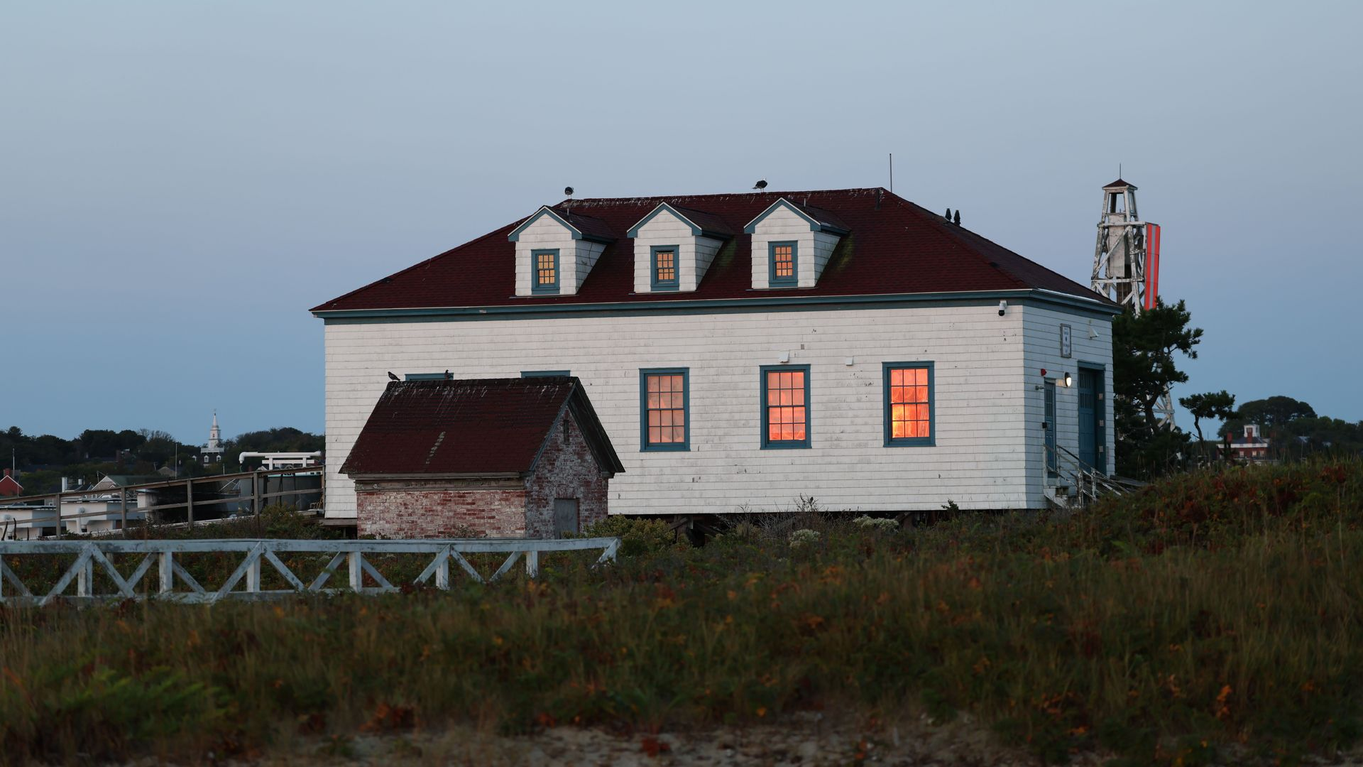 White lighthouse keeper’s house with lit windows at dusk on a grassy coastal hill